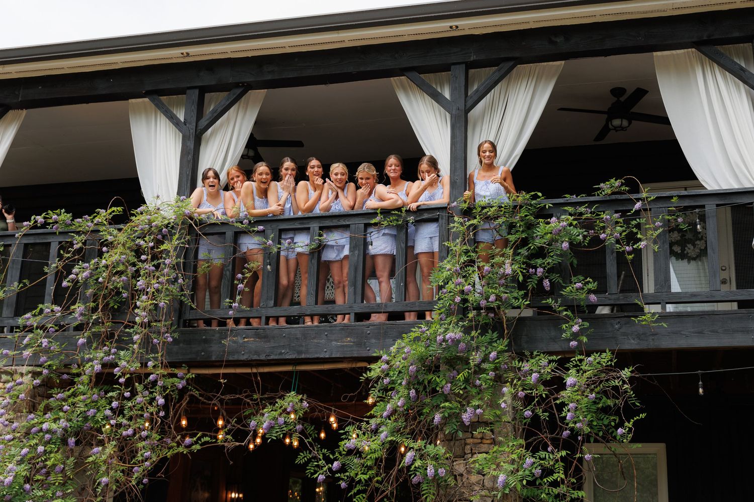 Group of women in matching outfits posing on a decorated balcony with lush greenery at an outdoor venue.