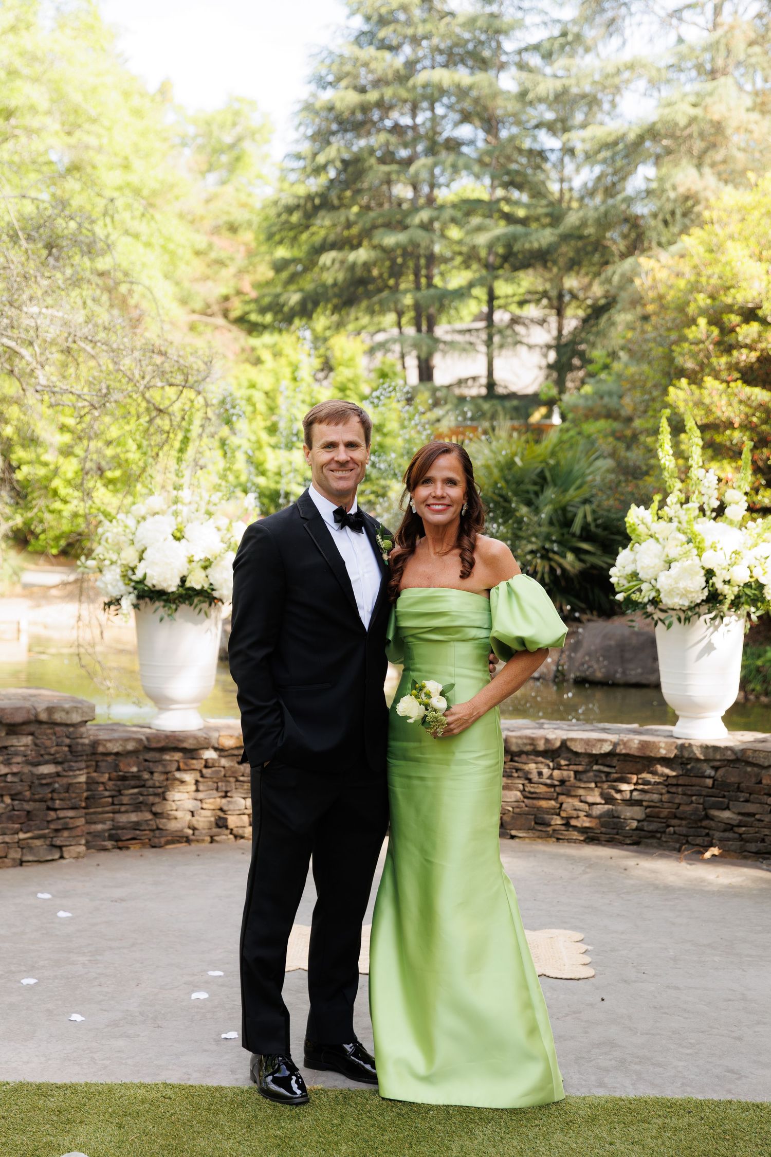 Couple posing outdoors at a formal event, man in tuxedo and woman in green off-shoulder gown, surrounded by white floral arrangements.