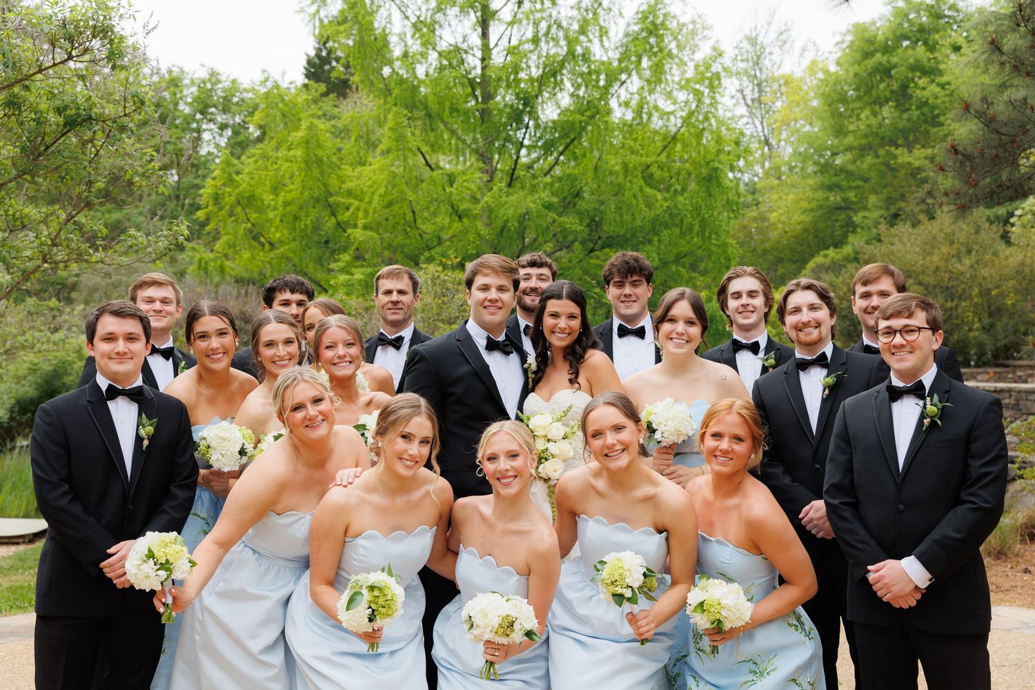 Large wedding party posing outdoors in formal attire, bridesmaids in light blue gowns holding white bouquets with groomsmen in tuxedos.
