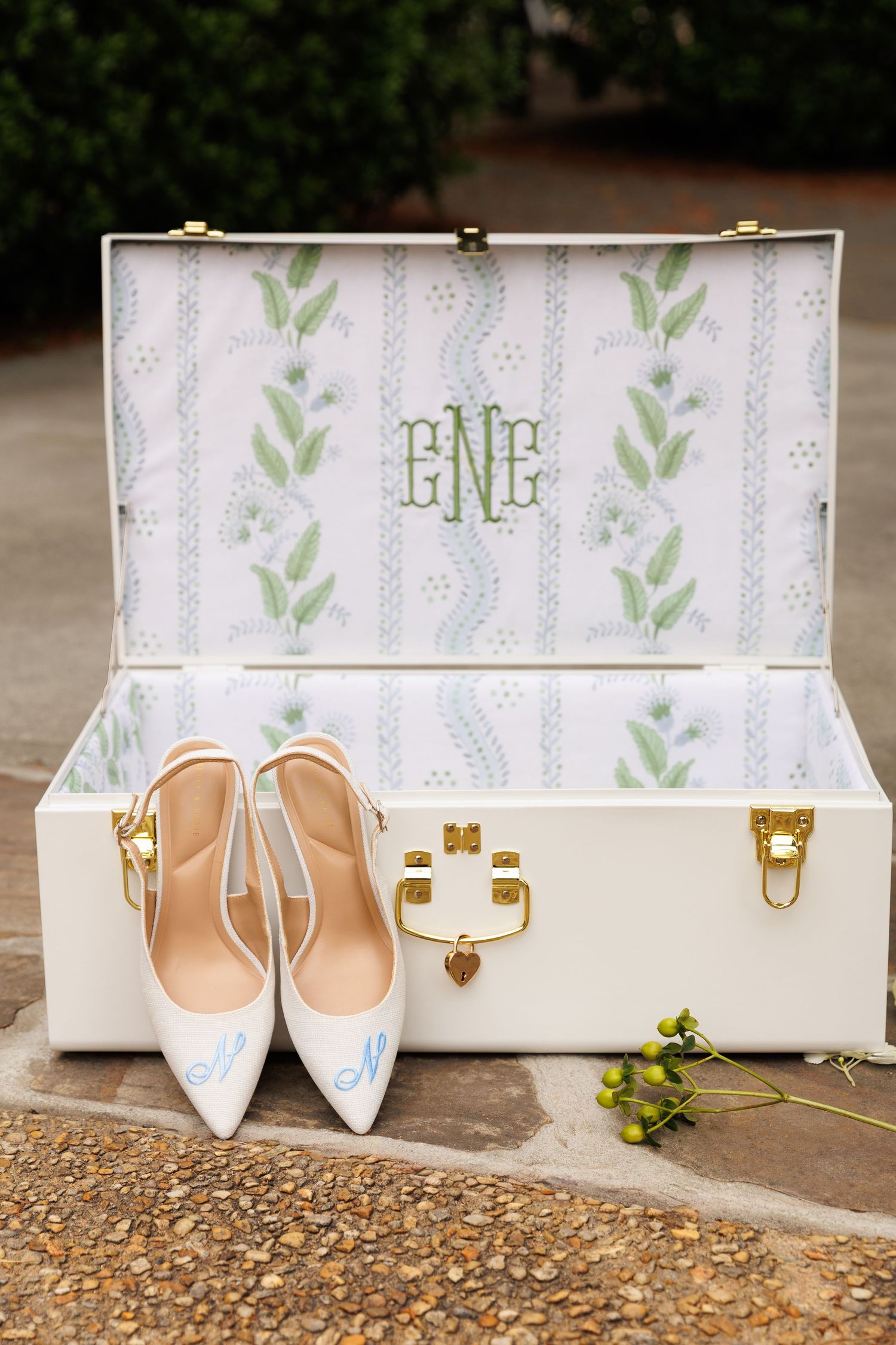 White monogrammed trunk with gold hardware displaying custom blue script bridal heels on stone patio.
