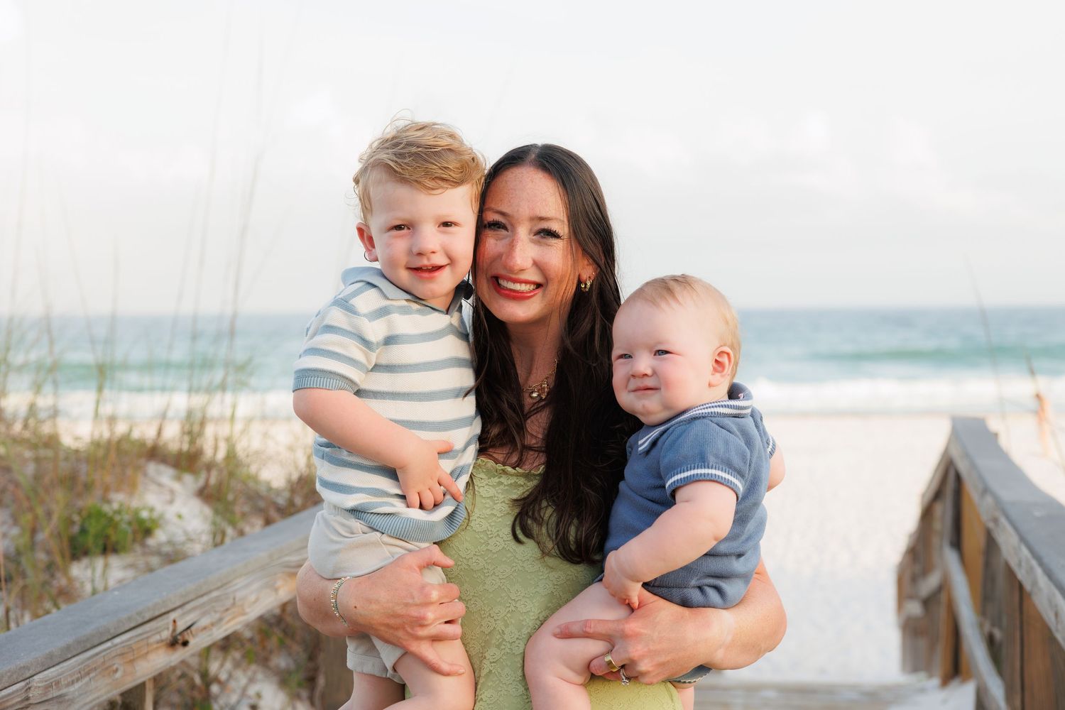 A smiling woman with dark hair holds two young boys on a sunny beach boardwalk with turquoise water behind them.