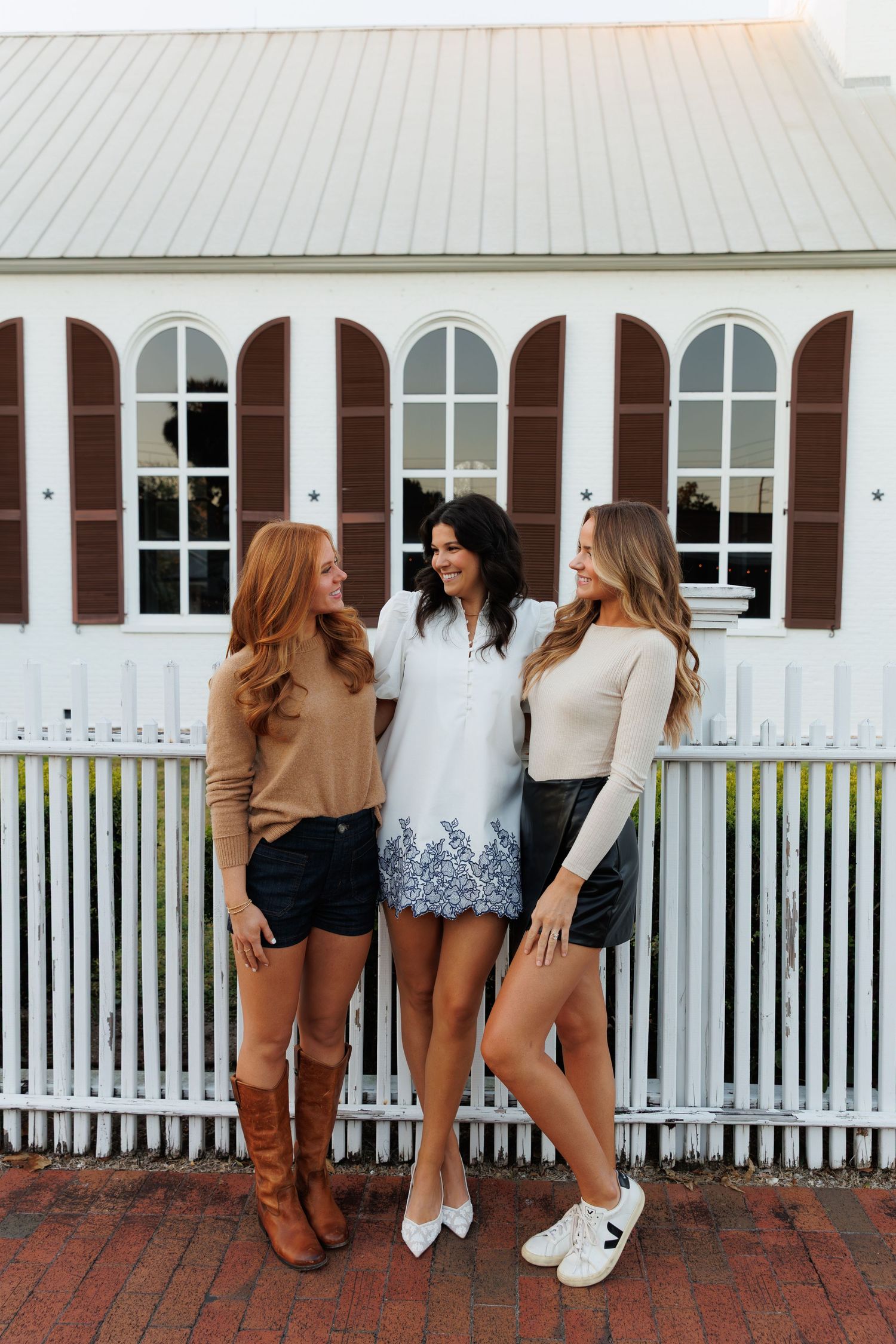 Three stylish women pose together in front of a white picket fence and building with arched windows.