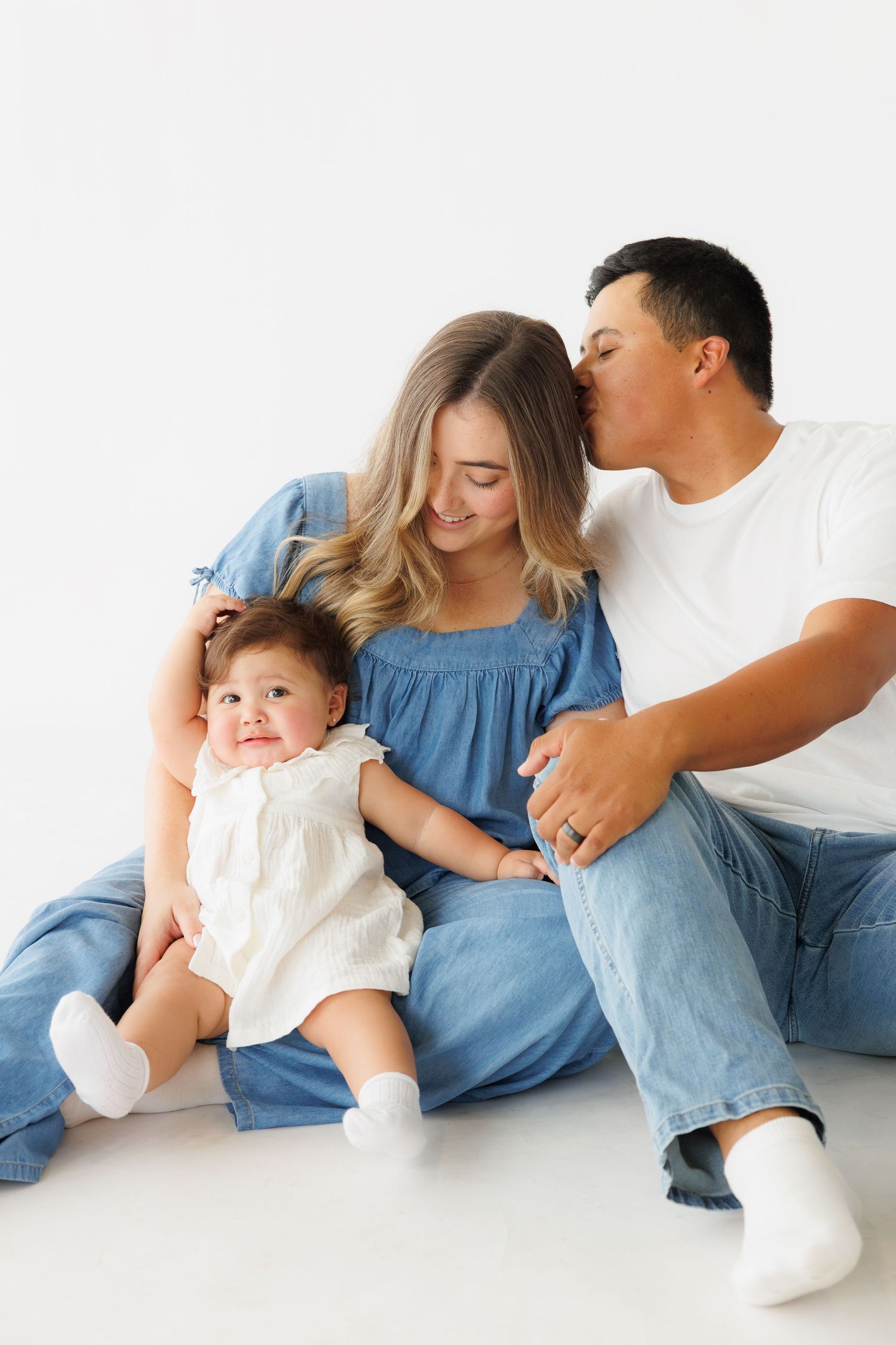 A happy young family with a baby girl in a white dress sits together on a white background for a studio portrait.