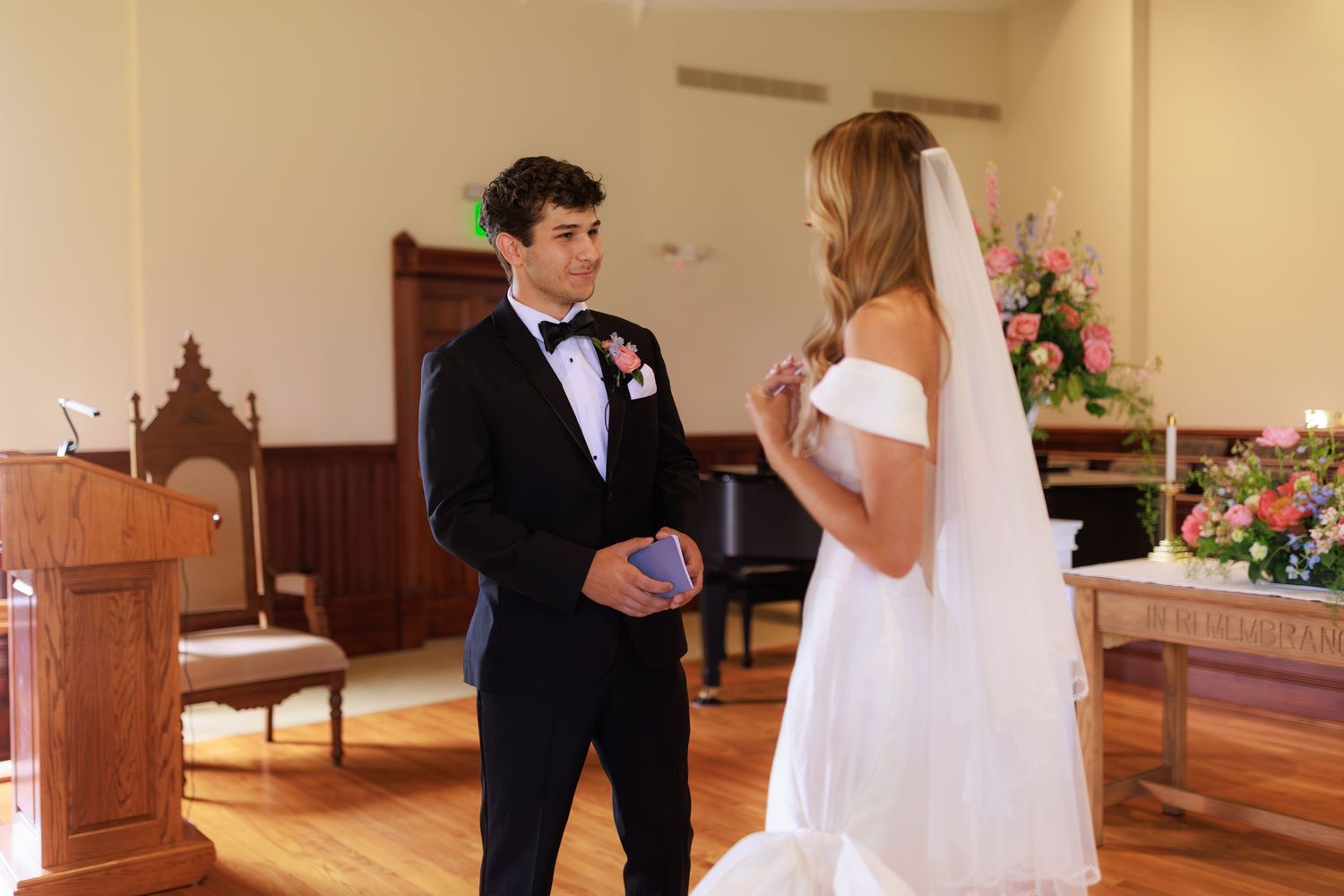 Two people in formal wedding attire share a moment during their ceremony in a church with floral decorations.