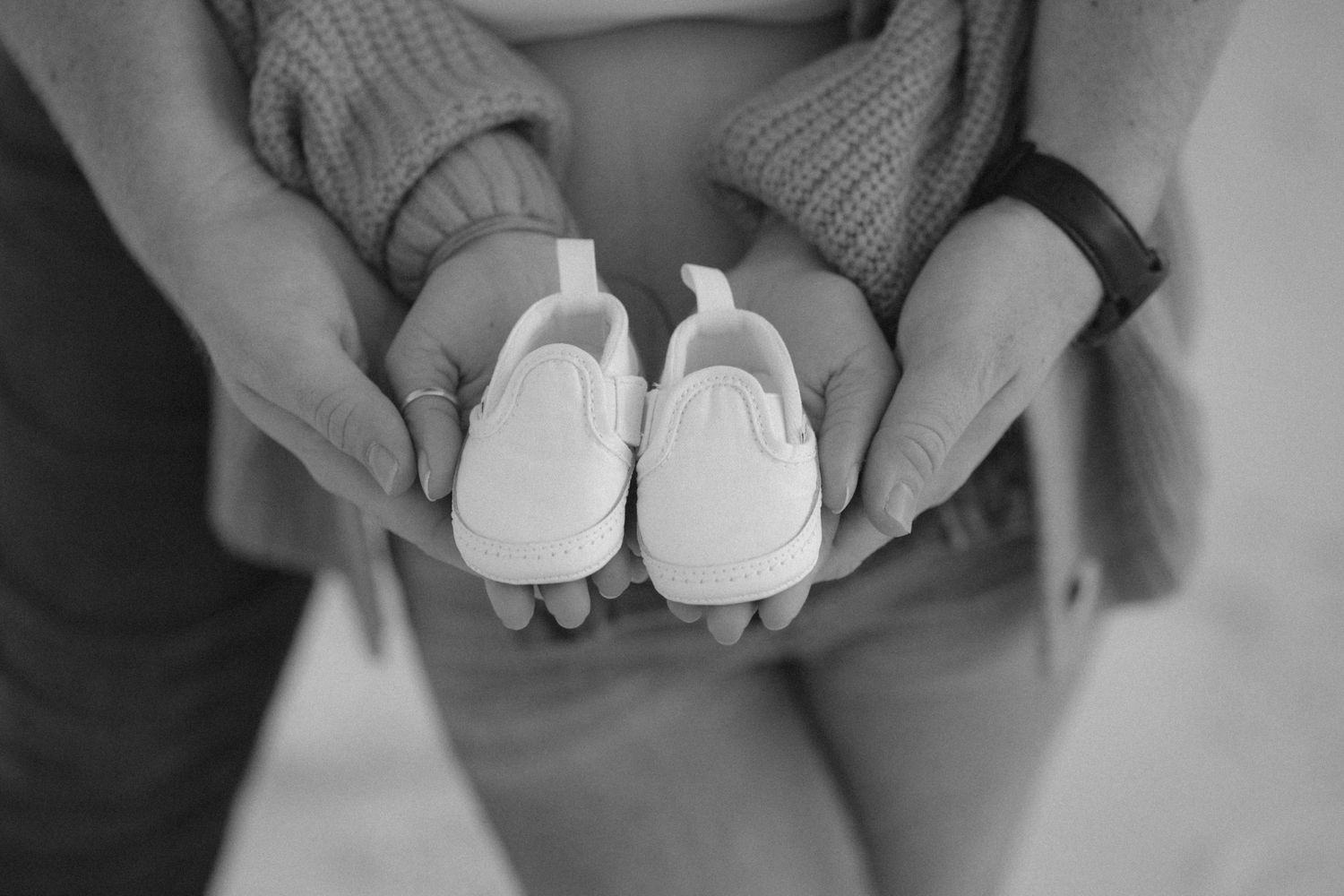 A pair of small white baby shoes being held in hands against a soft gray background.
