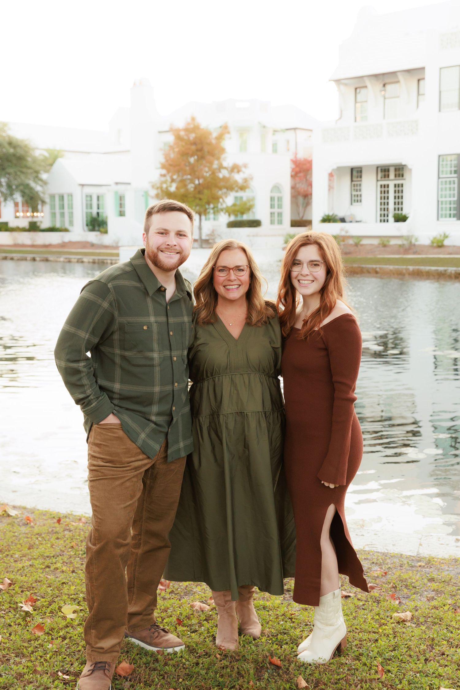 Three people pose together outdoors near a pond with white building and fall foliage in background.