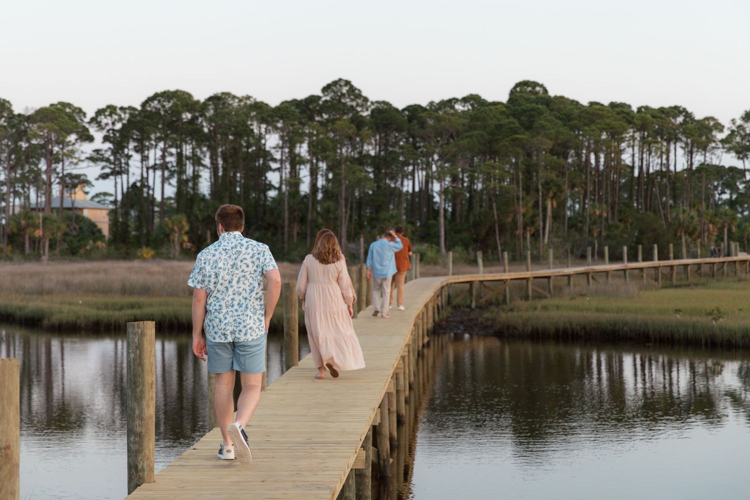 A group of people walk along a wooden dock overlooking a serene waterfront with pine trees in the background.
