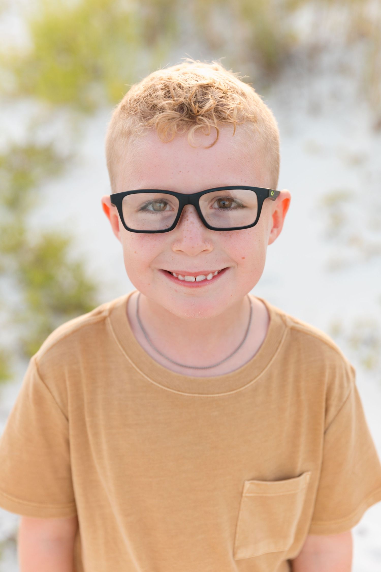 A close-up portrait of a young person wearing glasses and a tan t-shirt with a bright smile.