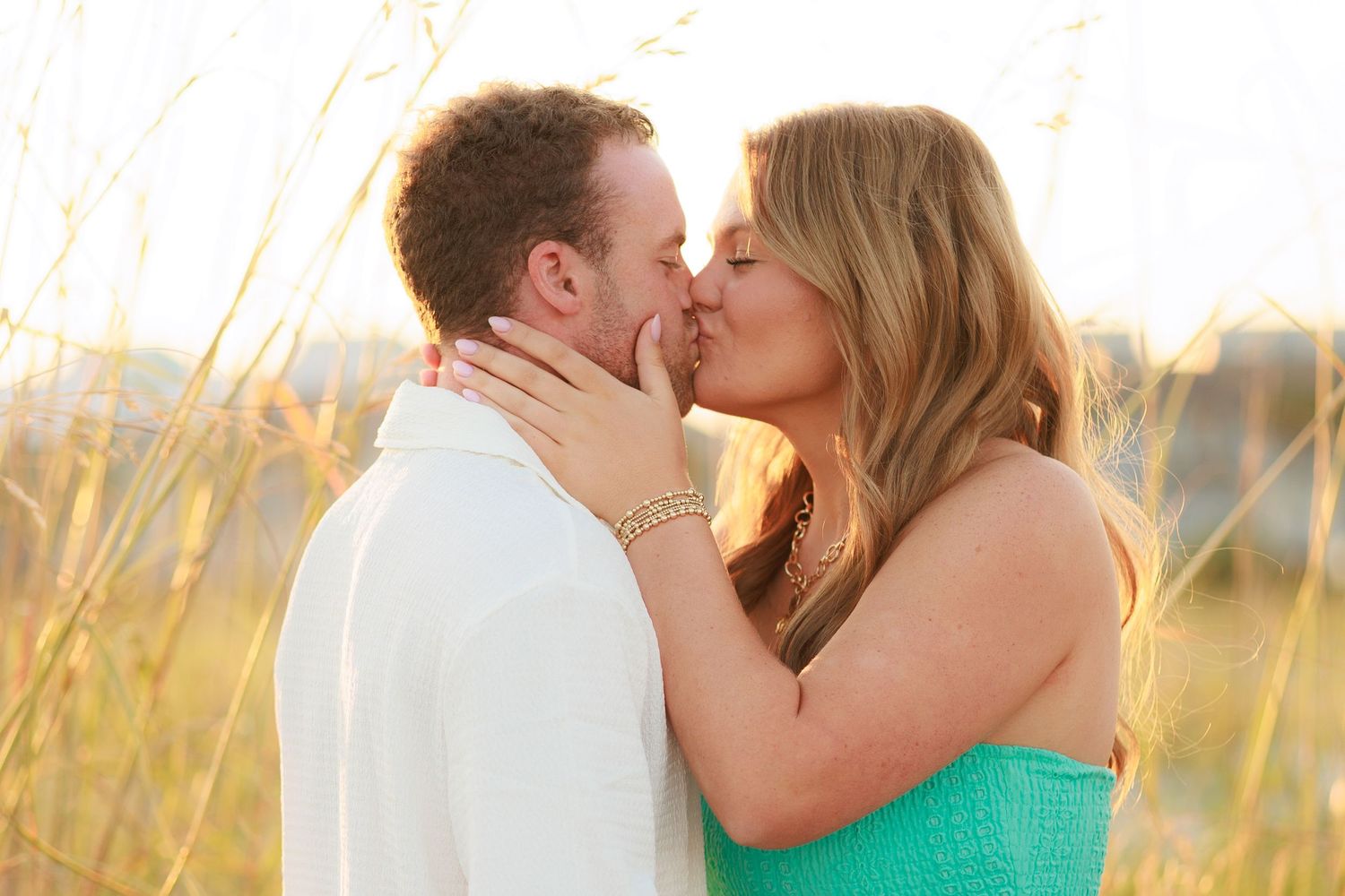 A romantic couple shares a tender kiss in a sunlit field of tall grass during golden hour.