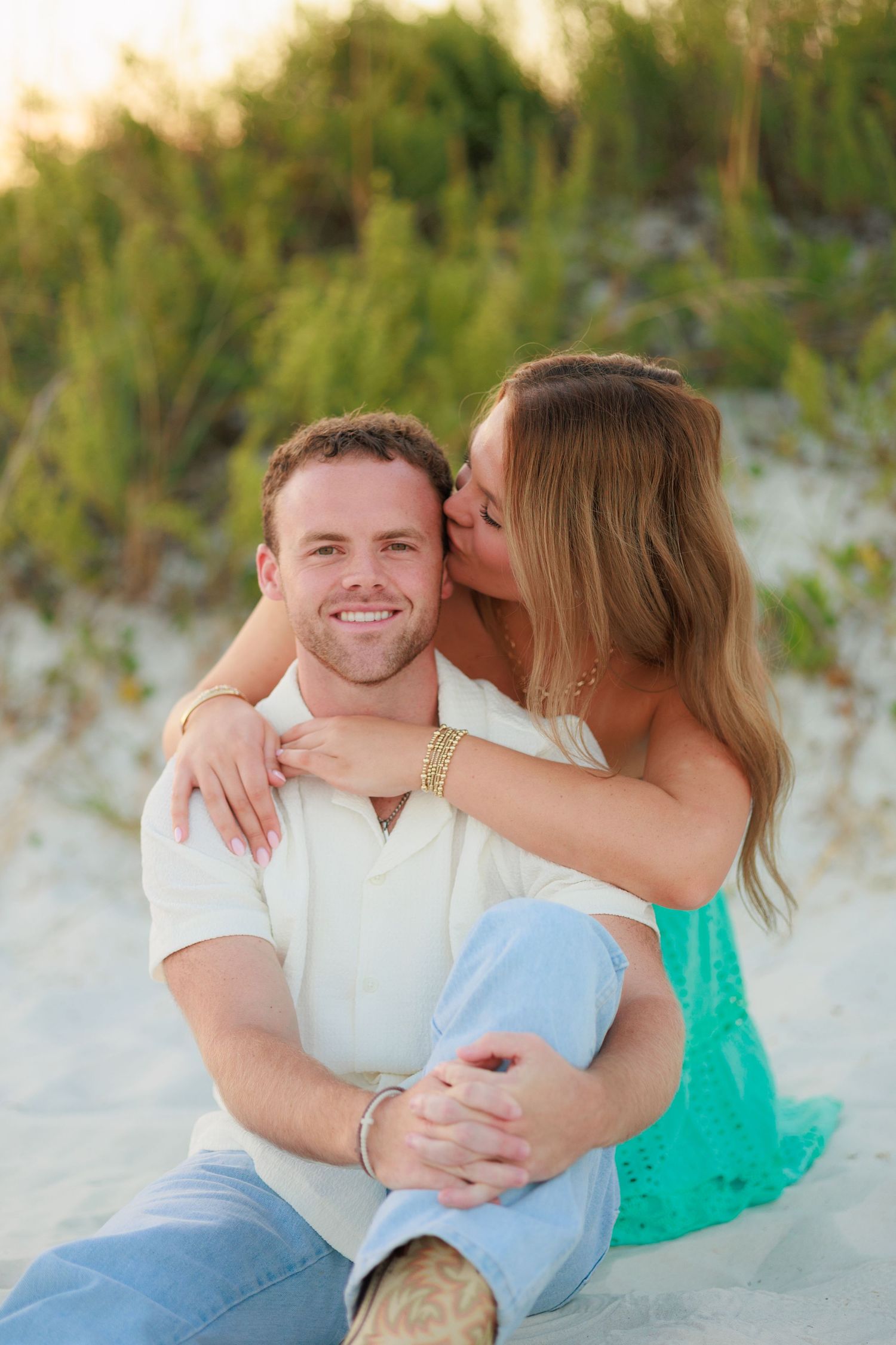A couple shares an intimate moment together on a sandy beach with green foliage in the background.