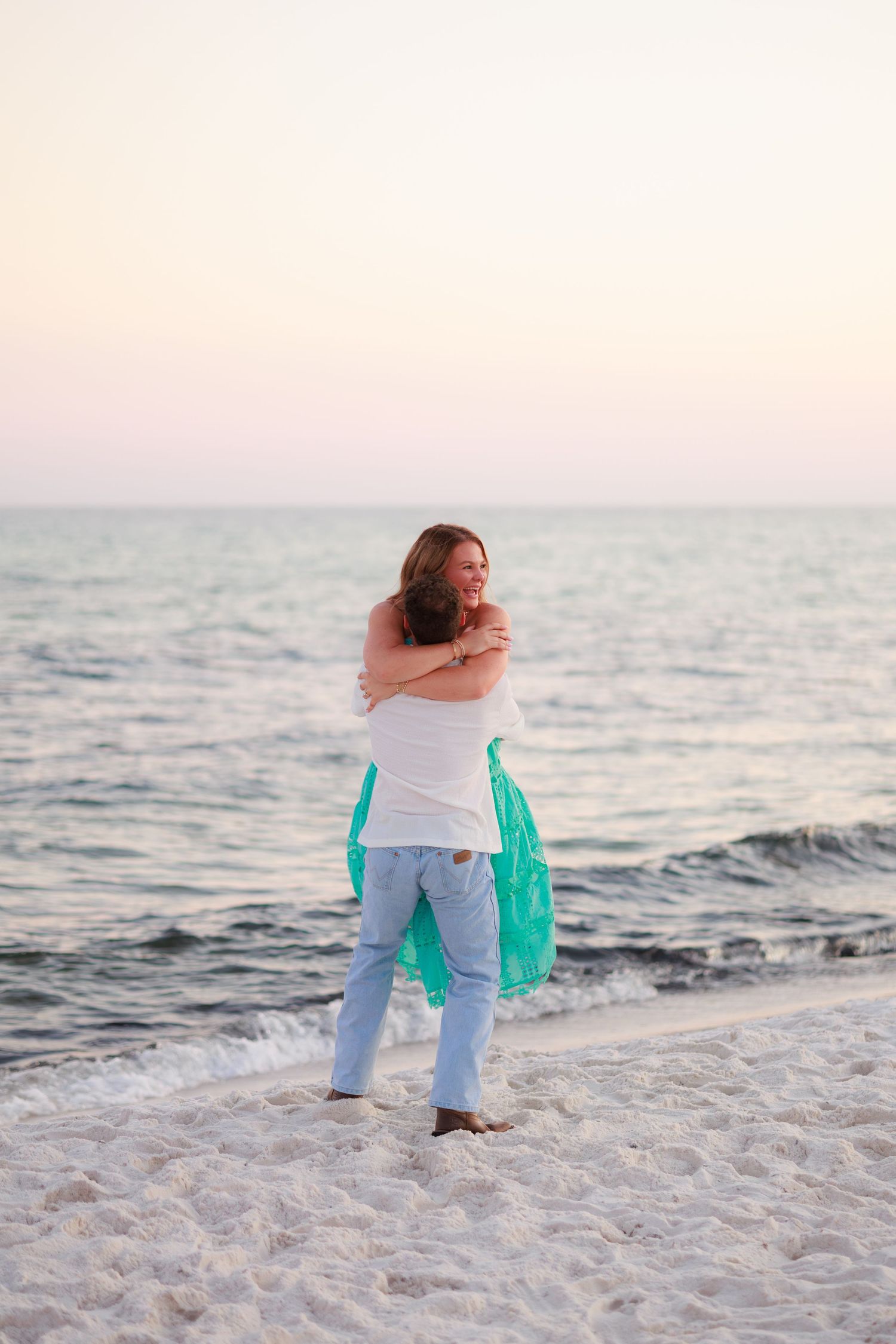 Two people embracing on a white sandy beach at sunset with gentle waves in the background.