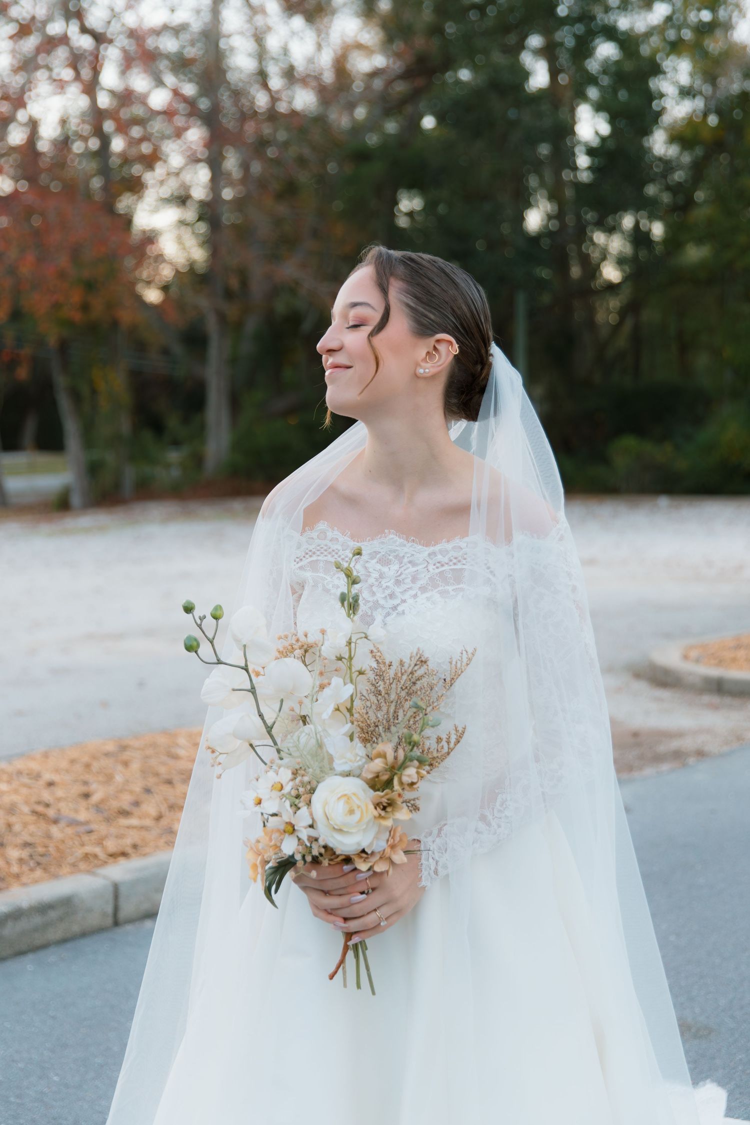 Bride in elegant white gown holding delicate bouquet against autumn backdrop.