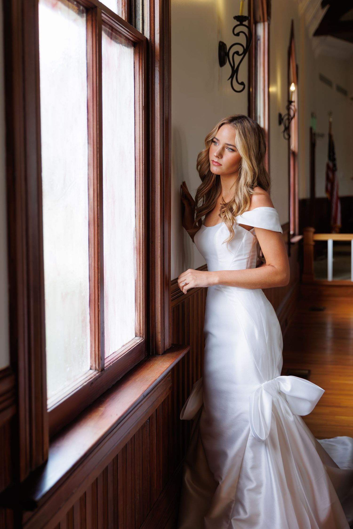 A bride in a white wedding dress gazes through a window in an elegant hallway with dark wood trim.