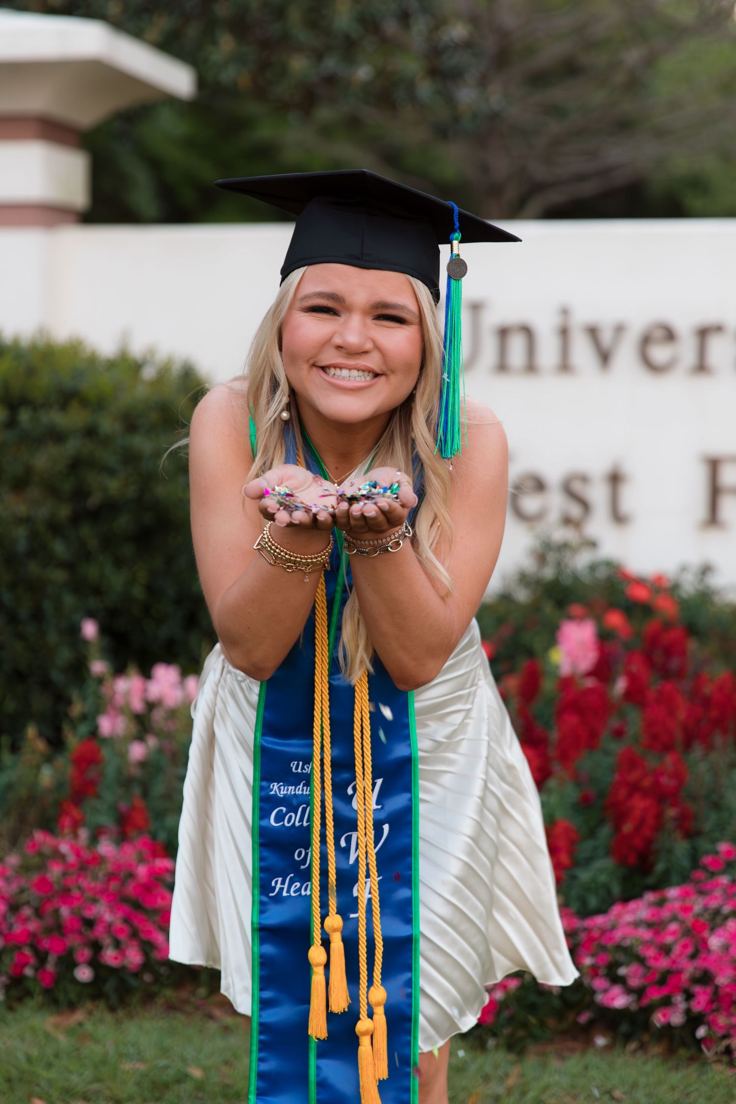 Graduate in cap and gown poses for photos with graduation stoles and pink flowers in the background.