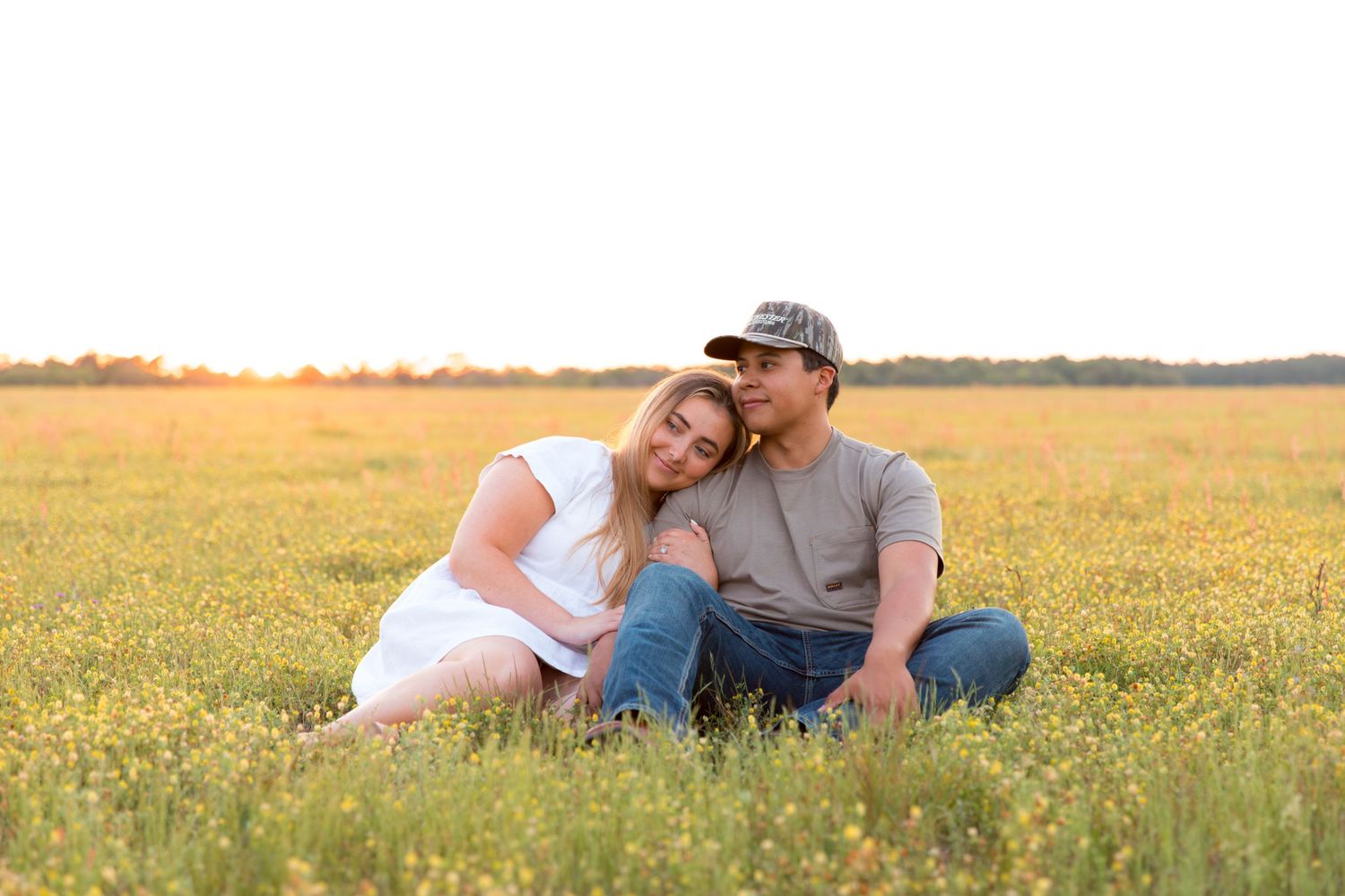Couple sitting together in a golden wildflower field at sunset, woman resting head on mans shoulder.
