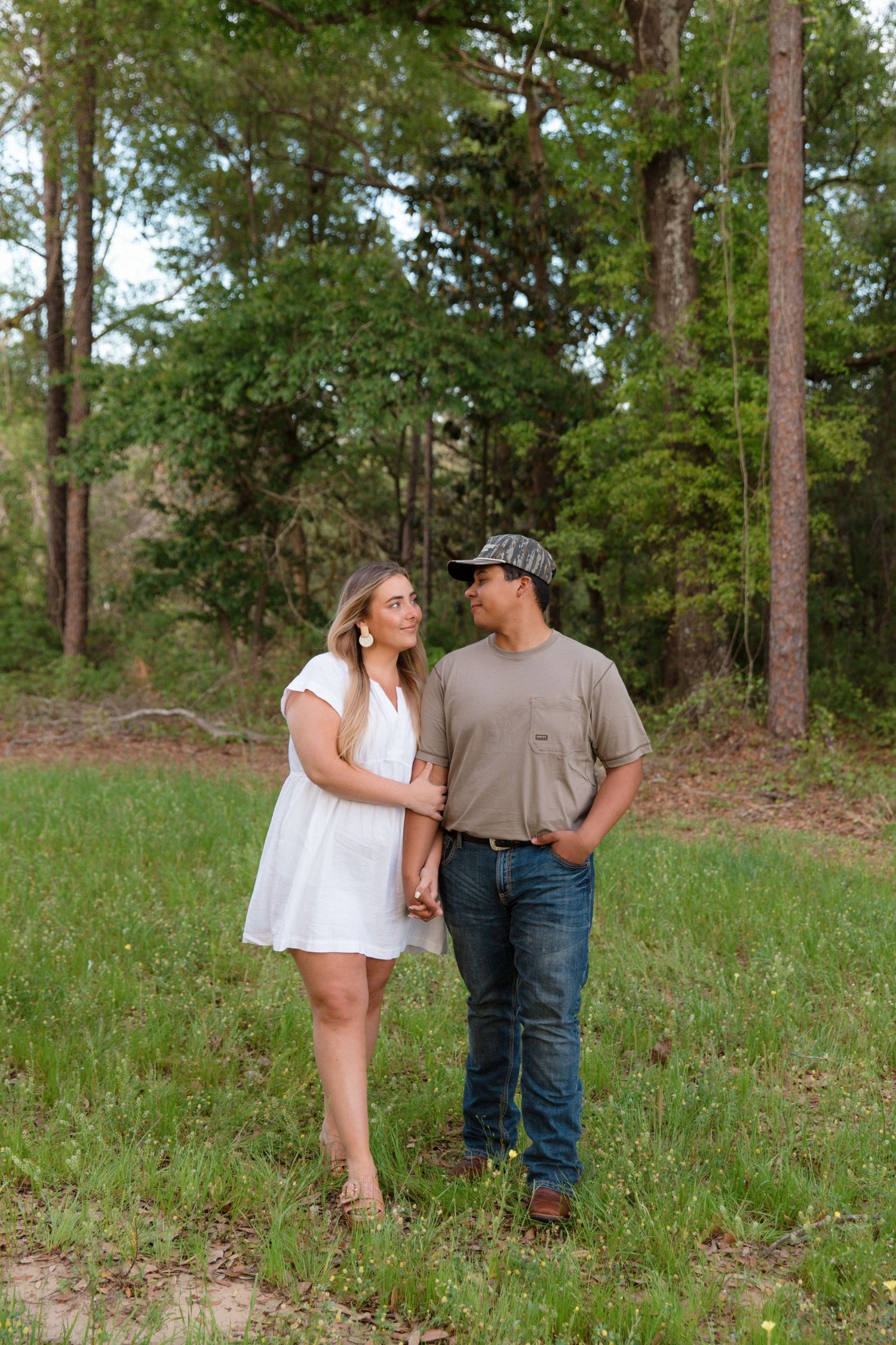 Couple holding hands in a green meadow with trees, woman in white dress and man in casual attire gazing at each other.