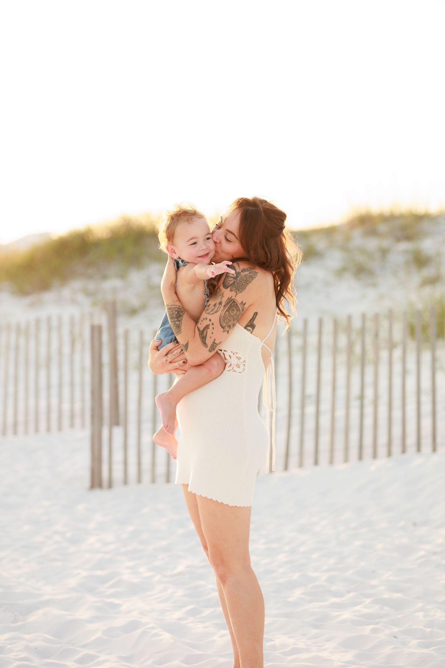 Tattooed mother in white dress kissing smiling baby on sandy beach with dunes and fence backdrop.