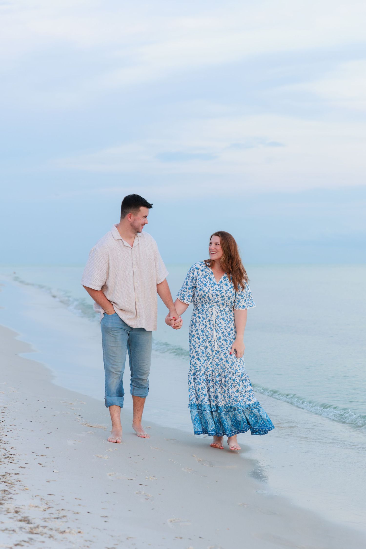A couple holds hands walking barefoot along a white sand beach at dusk, woman in blue floral dress.