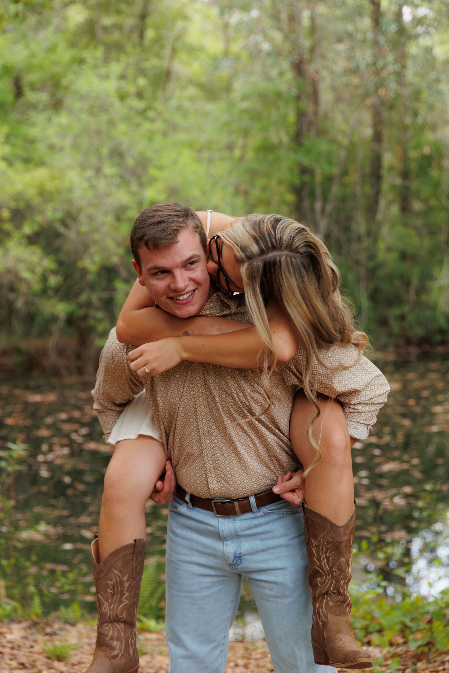 Laughing couple in a forest, man giving woman a piggyback ride, both wearing casual country-style clothing.