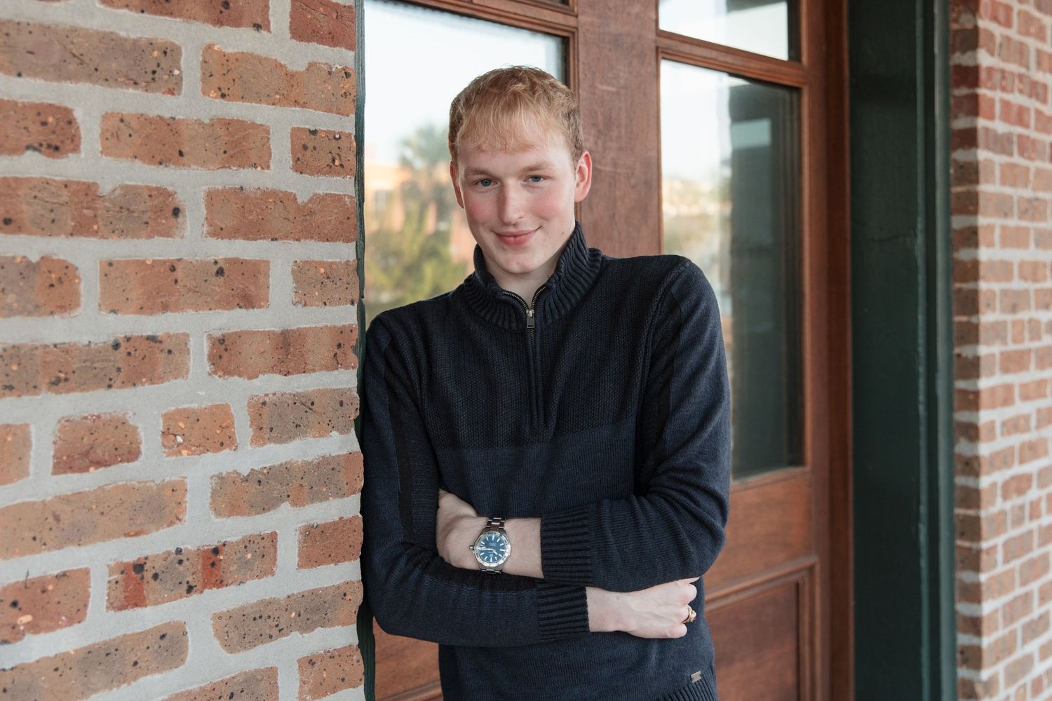 Young man in black zip-up sweater with arms crossed, smiling beside a brick wall and wooden door.