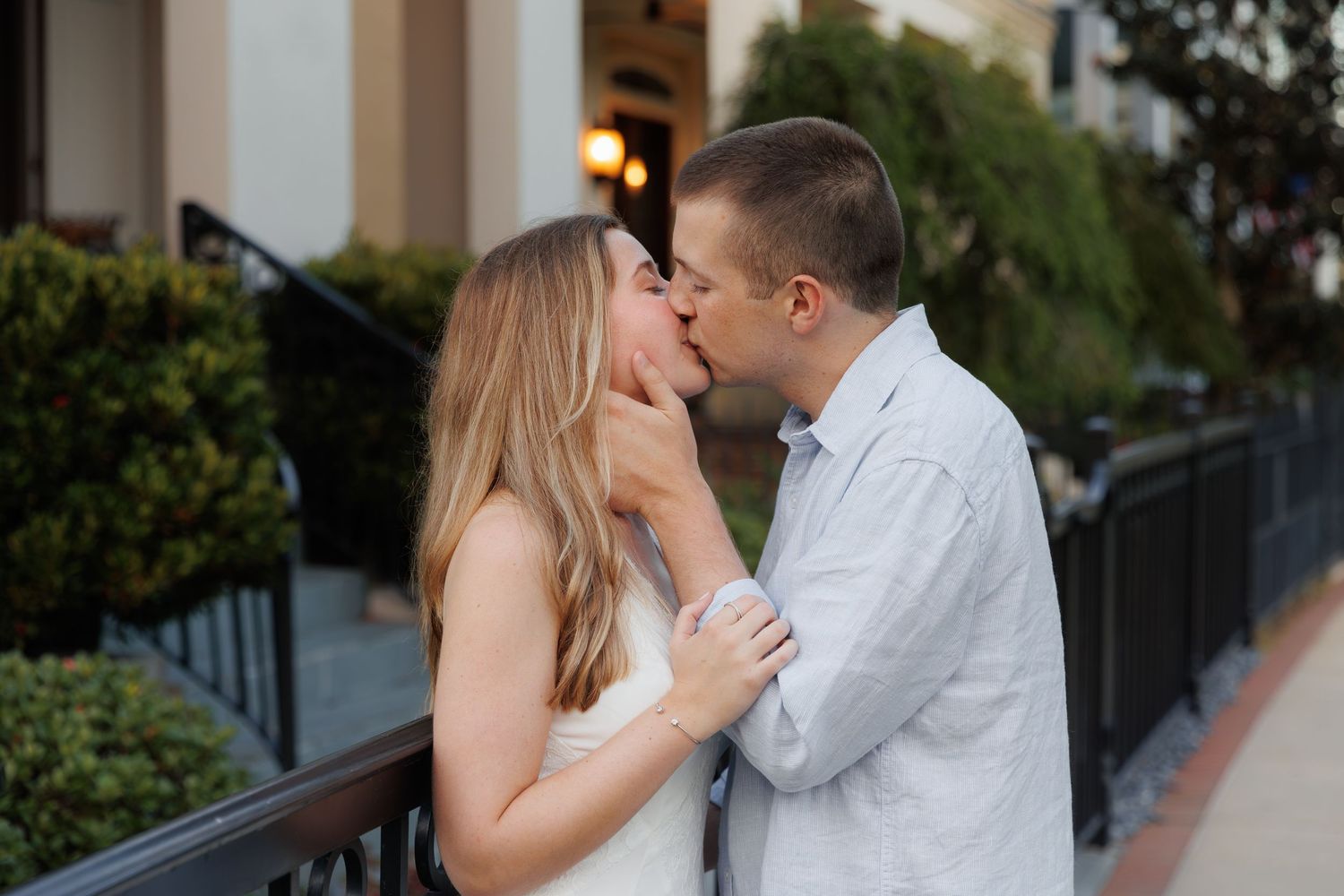 Couple sharing a romantic kiss outdoors near a railing, woman in white dress, man in light blue shirt.