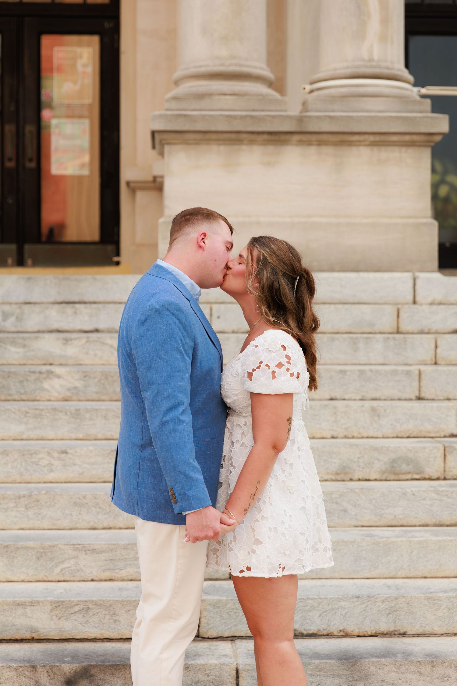 A couple shares a romantic kiss on stone steps outside a classical building, wearing a blue suit and white floral dress.