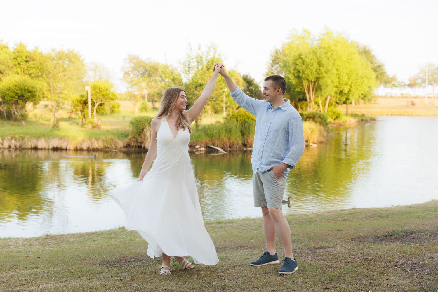 Couple dancing by a serene pond, woman in white dress, man in blue shirt and gray shorts, surrounded by lush greenery.