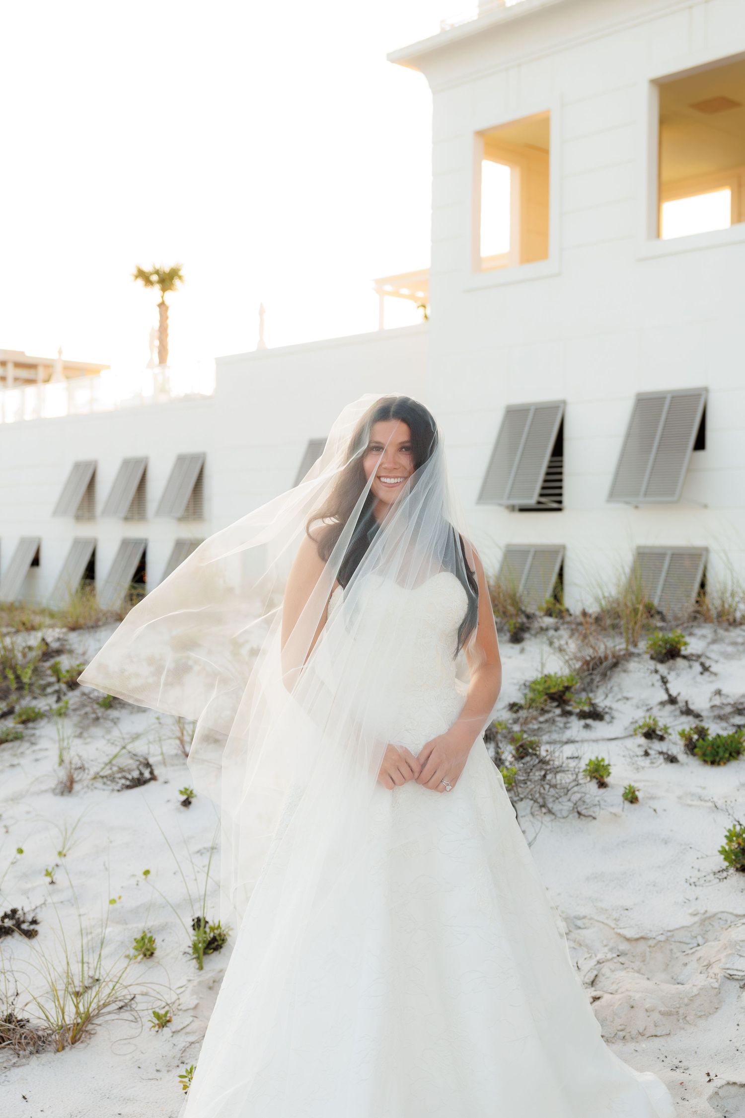 Bride in white ballgown with veil blowing in wind stands on sandy beach near white building.