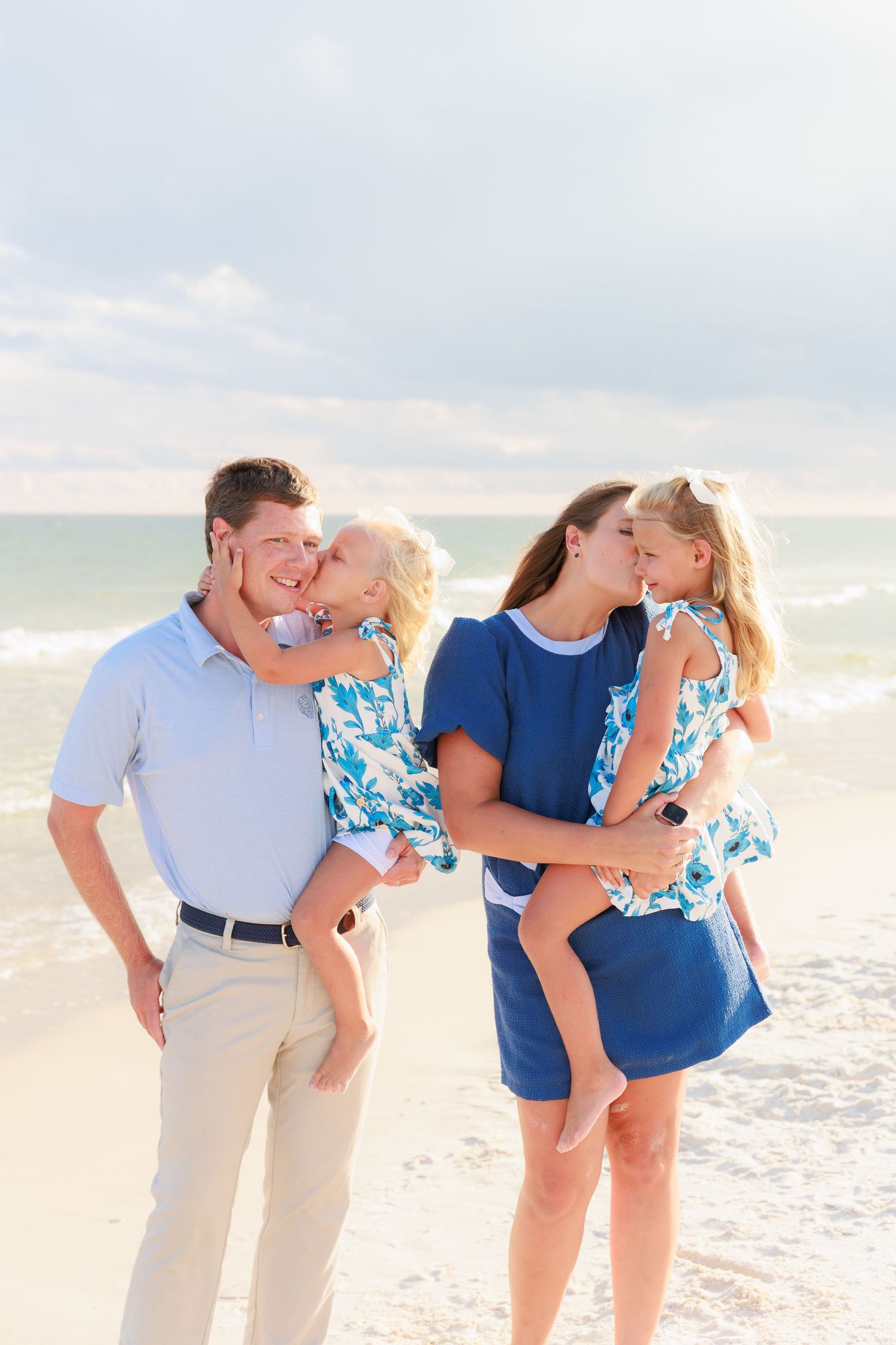 Happy family of four at the beach, two parents each holding a young girl who kisses their cheeks.