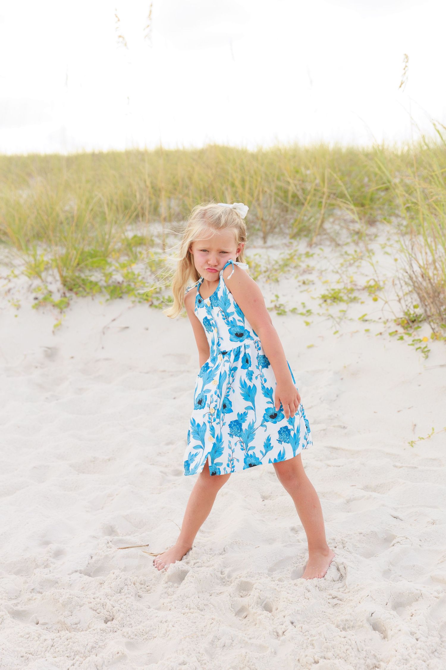 Young girl in blue floral dress playing on white sand beach dunes on a sunny day.