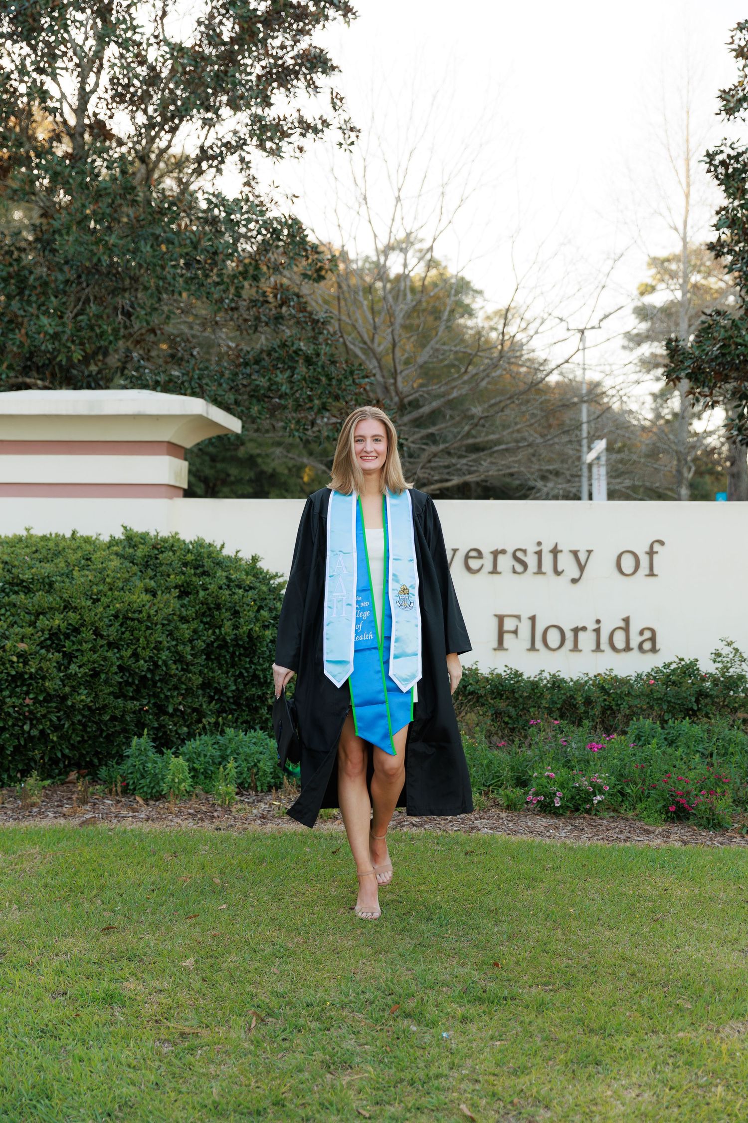 A graduate in cap and gown poses in front of the University of Florida entrance sign on a sunny day.