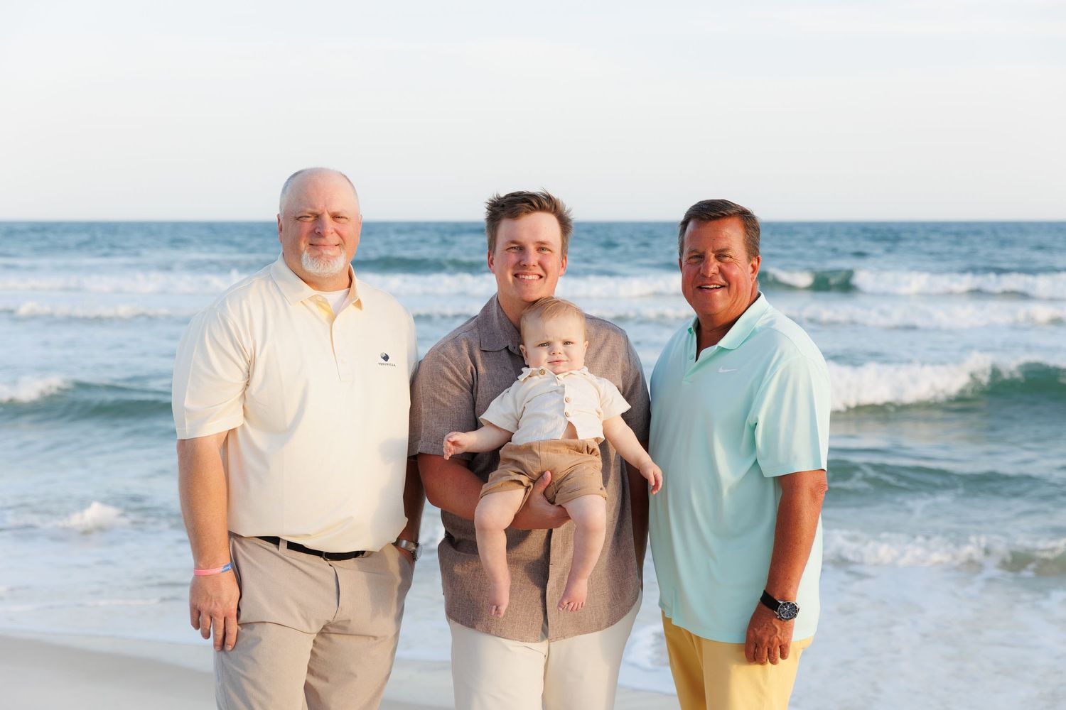 Three generations of men smile together on a sunny beach, with ocean waves in the background.