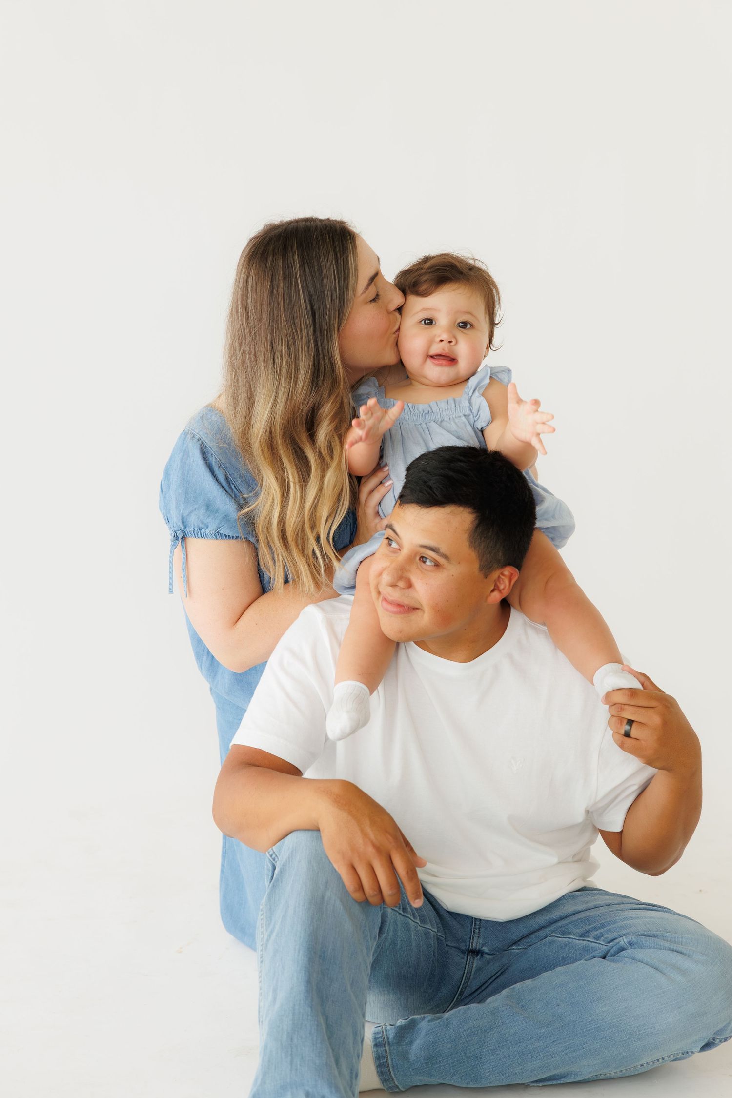 Mother kissing baby on fathers shoulders in white studio, sweet family portrait in denim outfits.
