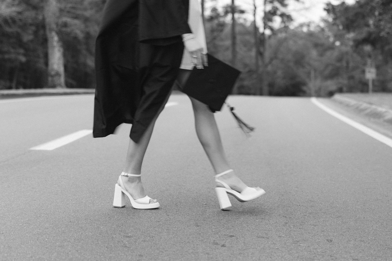 Black and white photo of a woman in heels and graduation gown walking on a road holding a diploma cap.