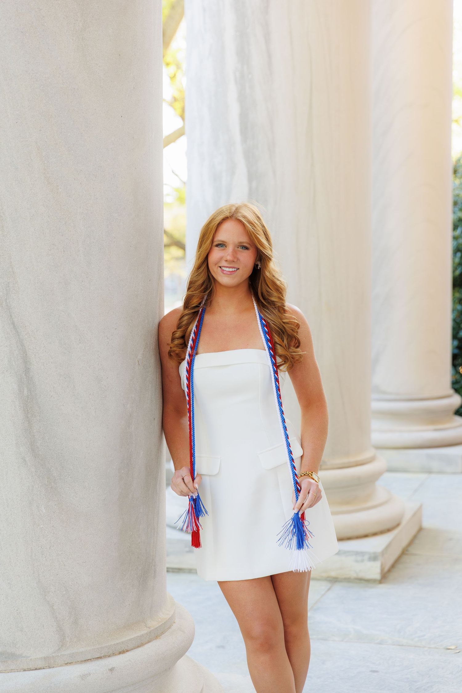 Young woman in white dress with red, white, and blue graduation cords posing between marble columns.