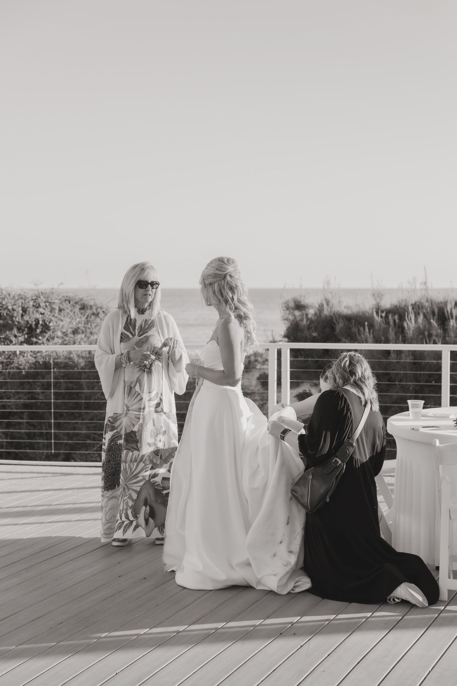 Black and white photo of a bride in a white gown with two women on a seaside deck at a wedding.