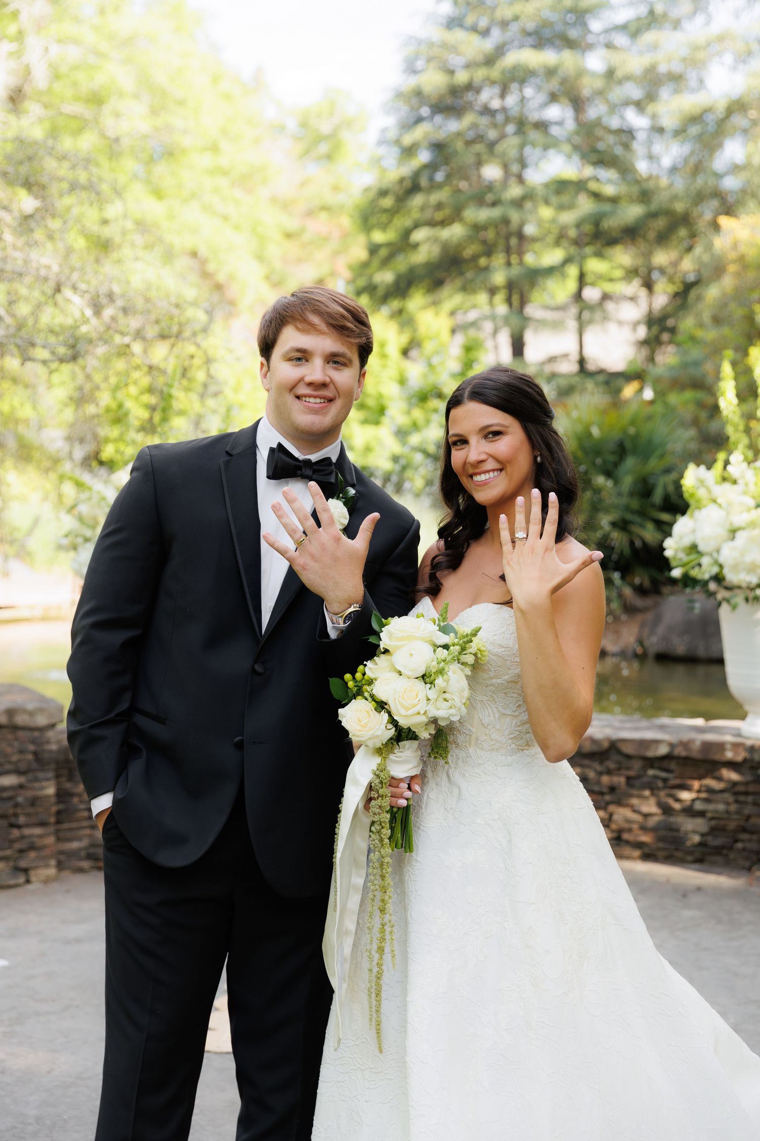 Newlywed couple smiling and showing wedding rings, groom in black tuxedo, bride in white gown holding white bouquet.