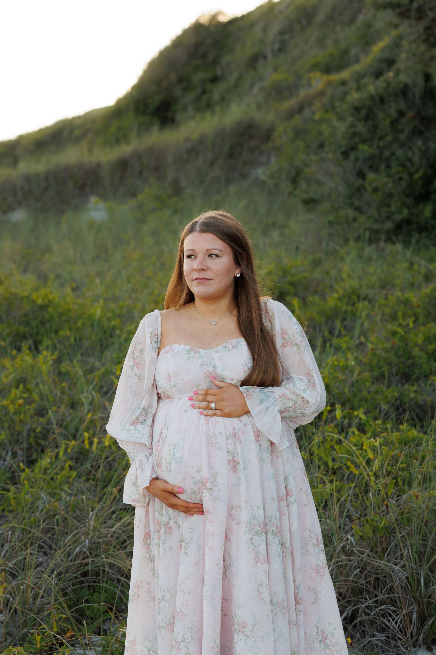 Pregnant woman in pink floral maxi dress cradles baby bump outdoors among lush green hillside.