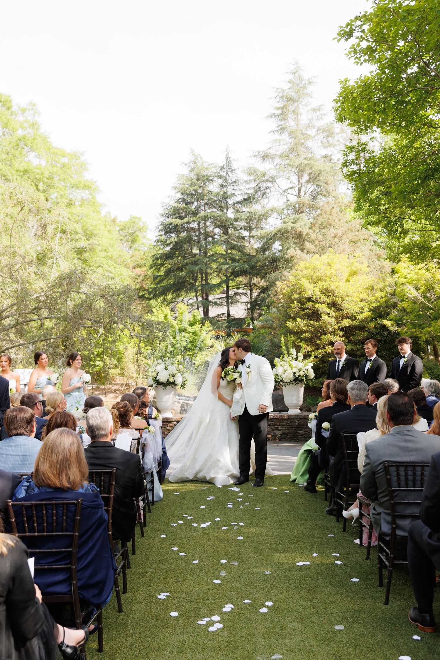 Bride and groom share first kiss at outdoor garden wedding ceremony with seated guests lining the petal-strewn aisle.