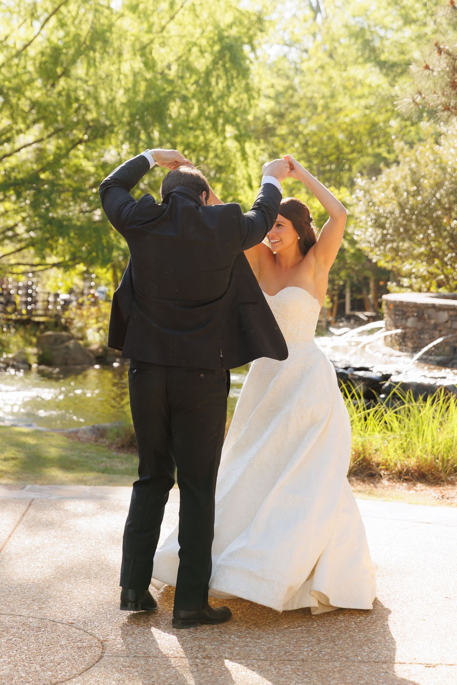 Bride and groom dance together outdoors, raising their arms joyfully near a scenic garden pond.