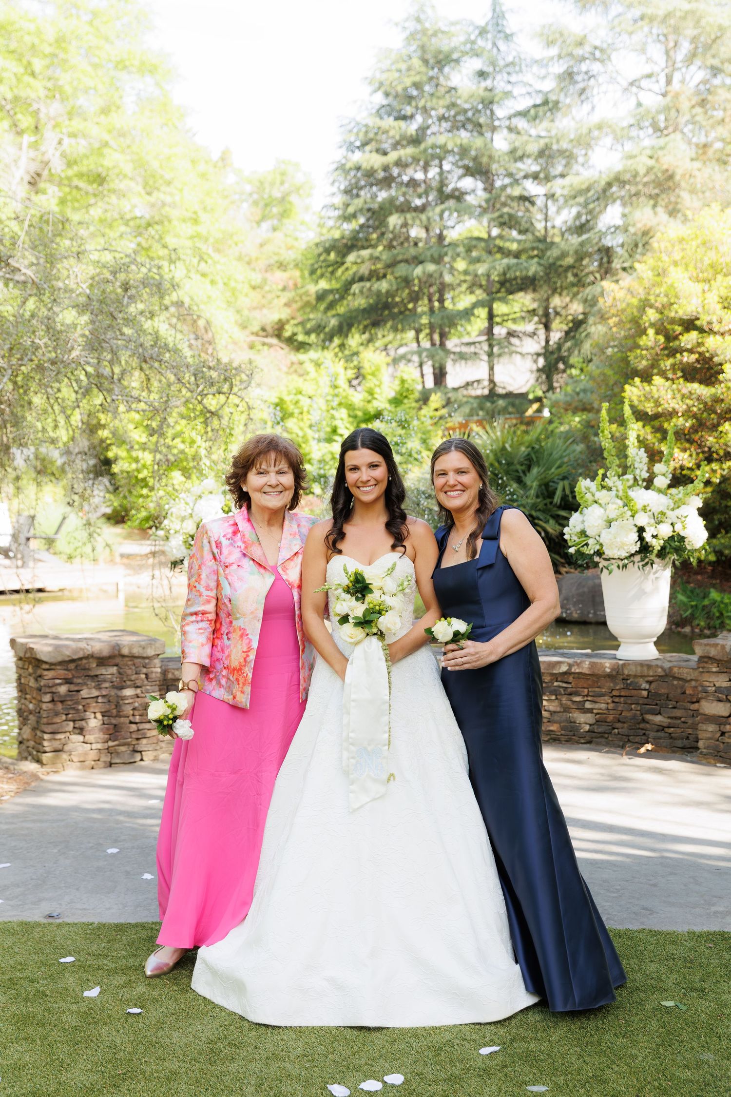 Bride in white gown poses with two women in pink and navy dresses, holding bouquet outdoors at a sunny garden wedding.