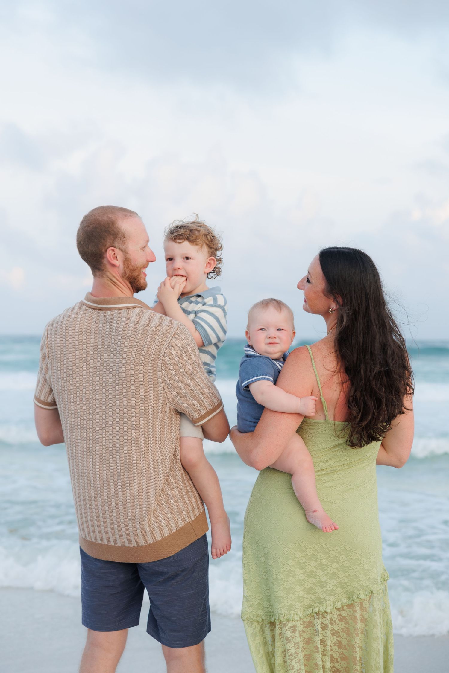 Family of four enjoying a beach day, parents holding two young children with ocean waves in the background.