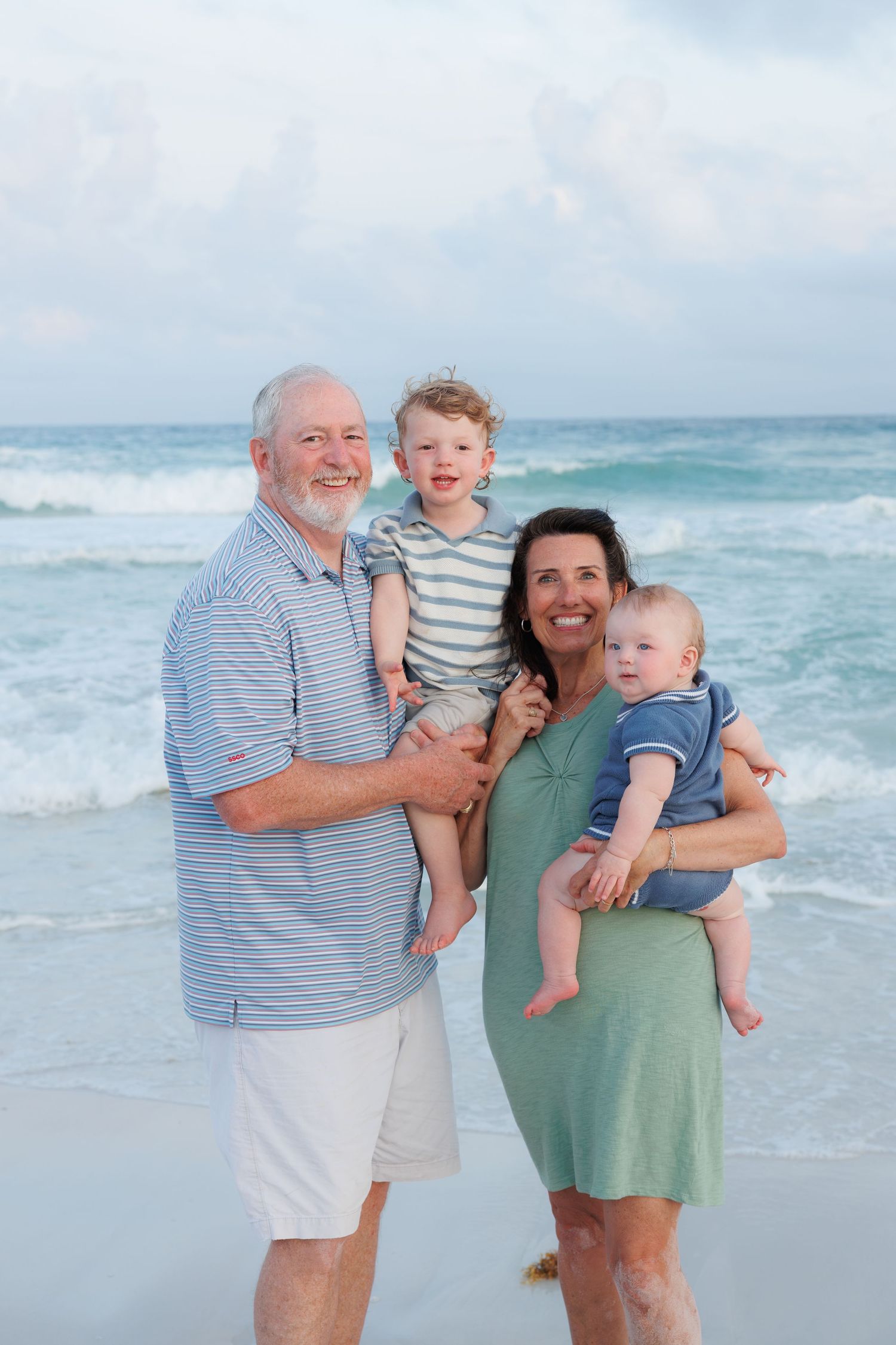 Grandparents holding two young grandchildren smiling on beach with ocean waves.