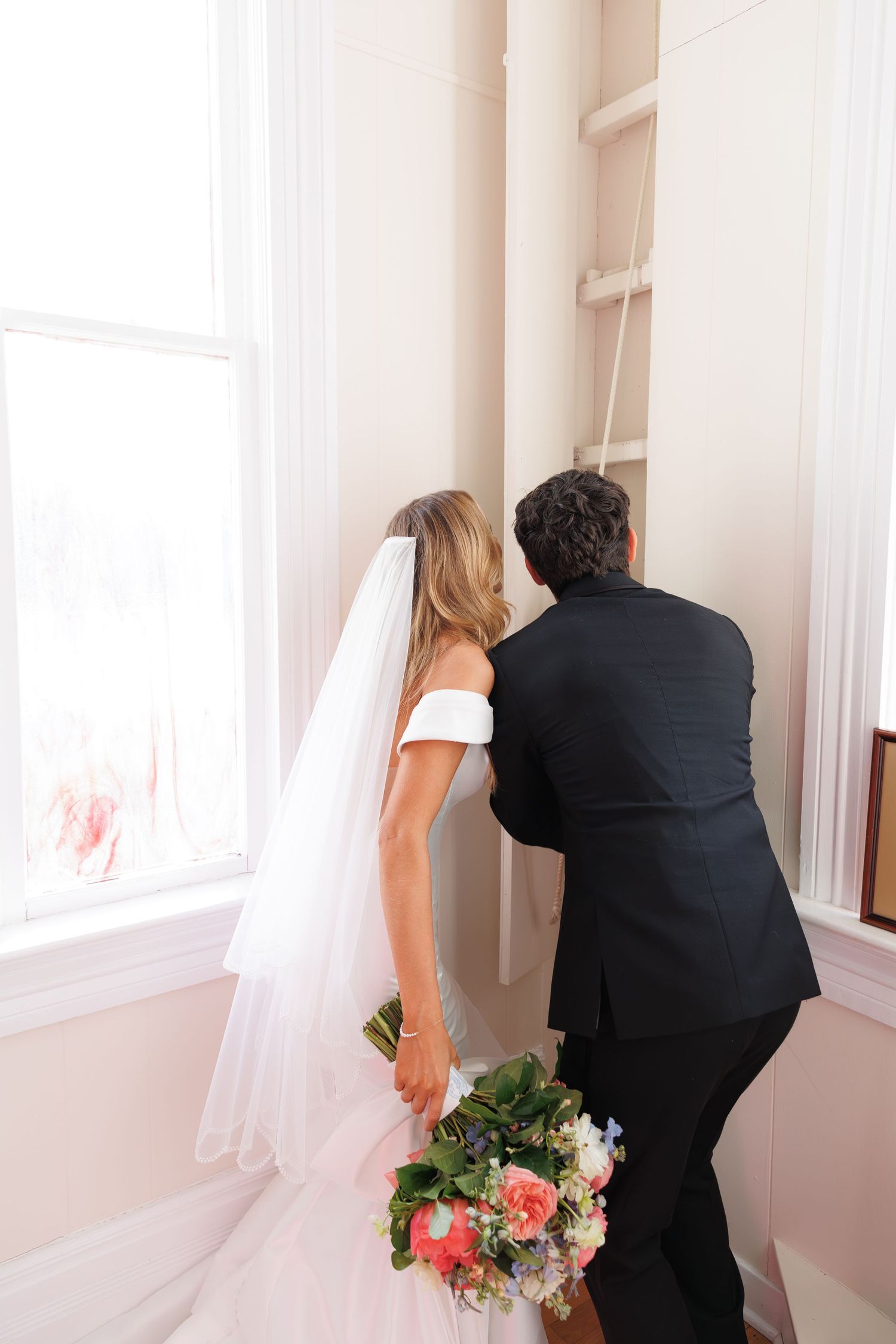 Bride in off-shoulder gown holds bouquet while looking out bright window with companion in formal attire.