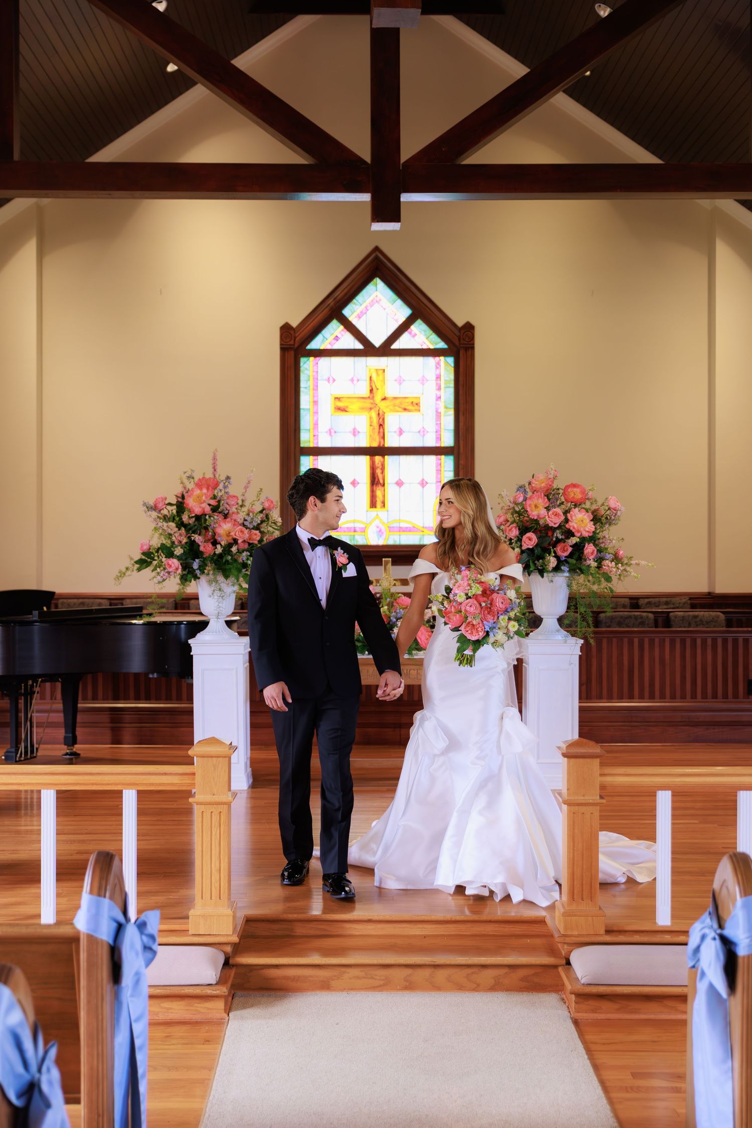 Newlywed couple walking down church aisle with stained glass cross window and pink floral arrangements.