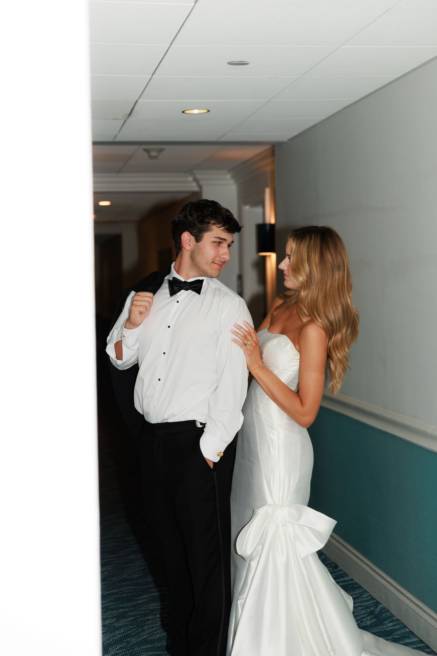 A couple in formalwear shares an intimate moment in a hotel hallway, the bride wearing a fitted white gown with bow detail.