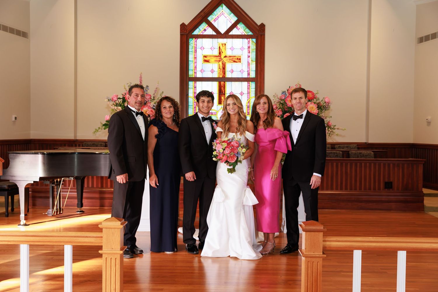 A wedding party poses together in front of a church altar with a cross window and wooden pews.
