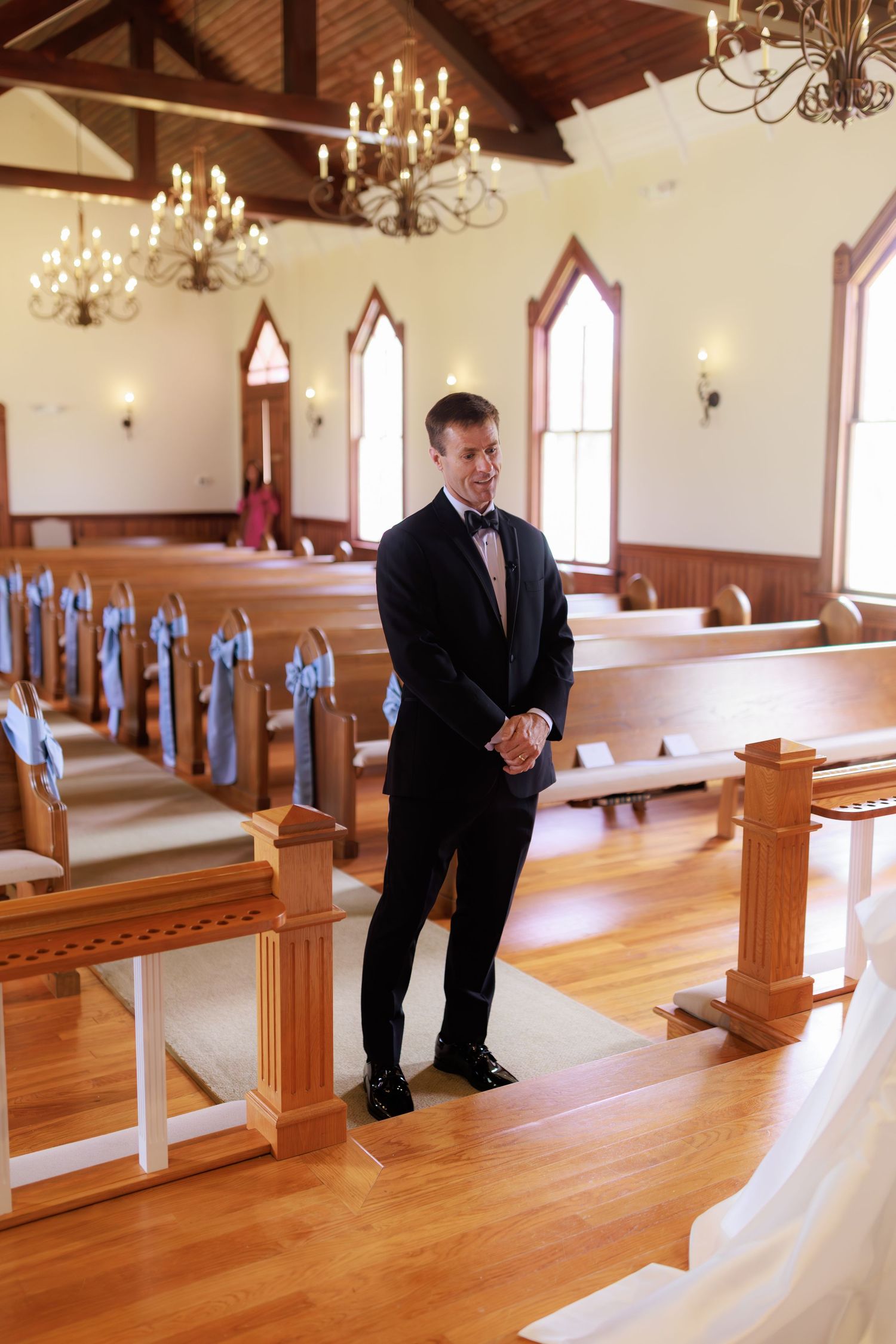Person in black suit bends down in wooden chapel interior with rows of pews and hanging chandeliers.