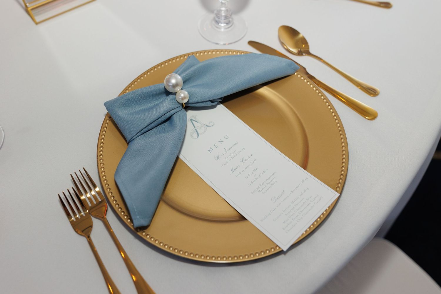 Elegant place setting with gold charger plate, blue napkin, menu card, and metallic gold utensils on white tablecloth.