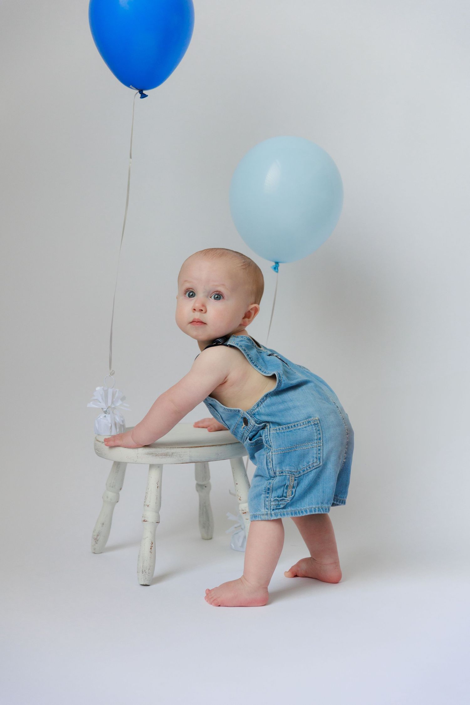 Baby in denim romper plays with white wooden chair while blue and light blue balloons float overhead.